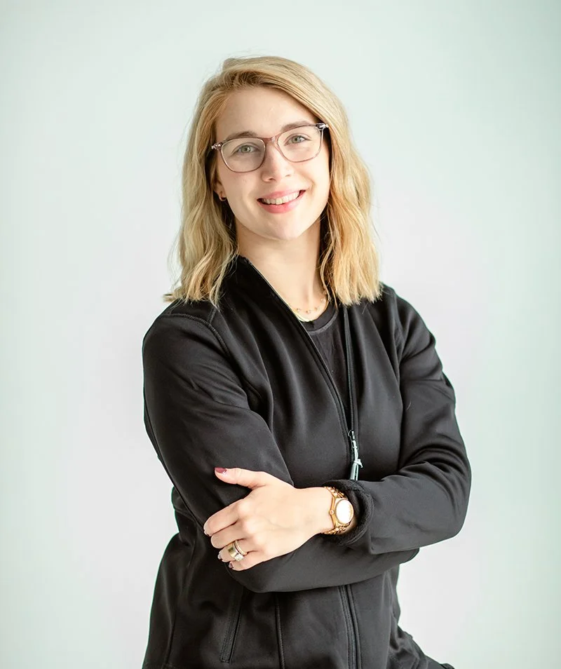 A young woman with blonde hair and glasses smiling, wearing a black jacket and standing with arms crossed against a light-colored background.