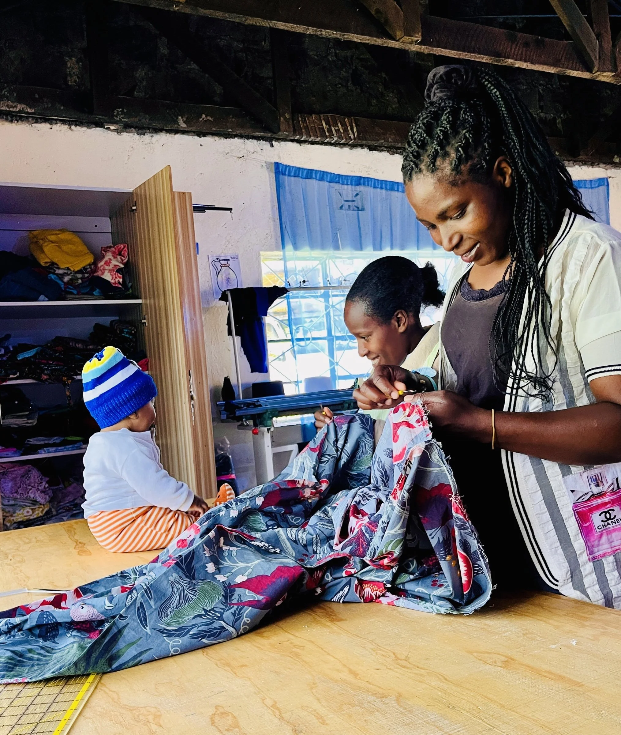 A woman and a girl smile as they work with floral fabric at a table in the Threads of Hope workshop, while a toddler in a knit cap sits nearby