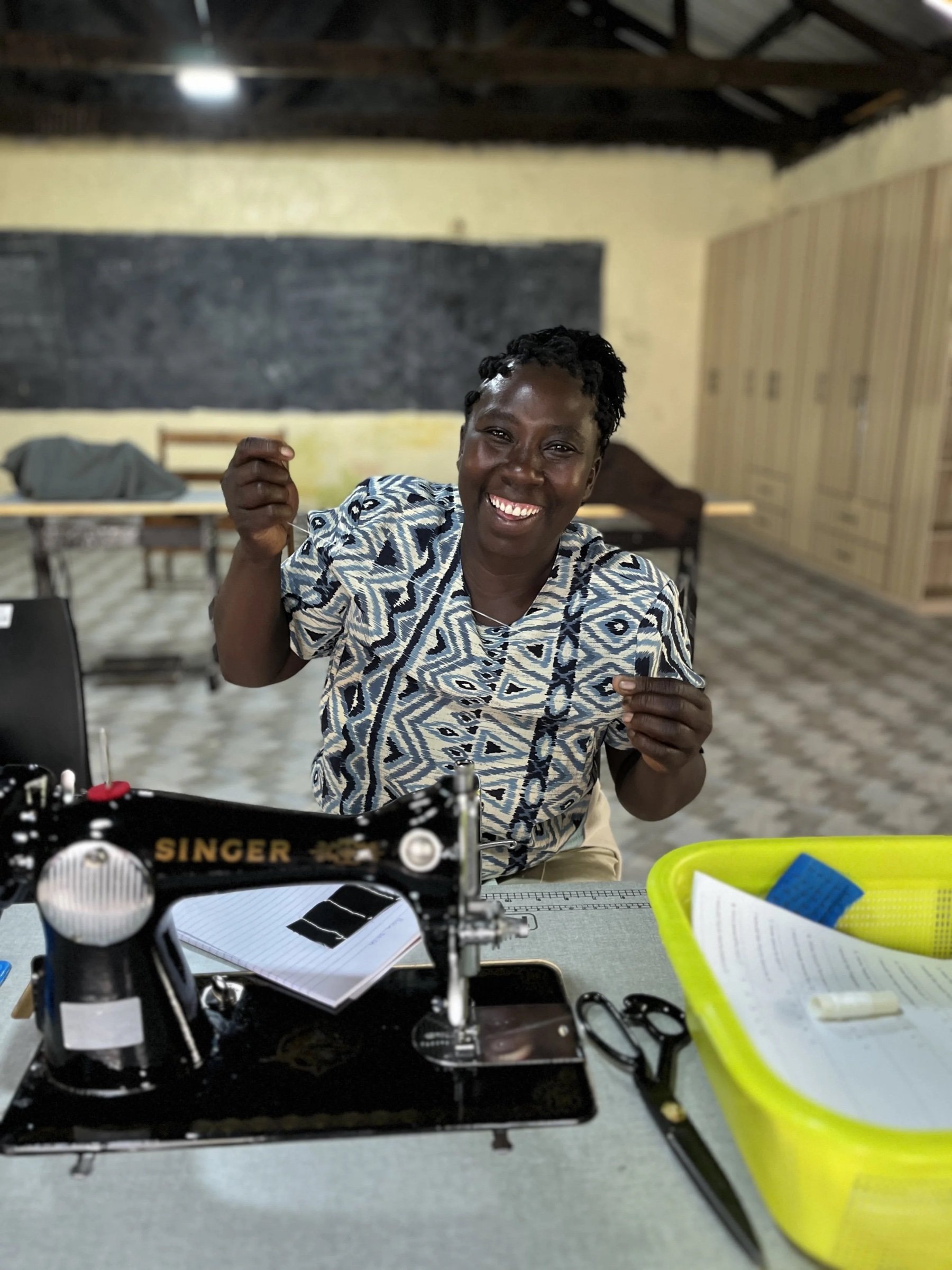 Loice smiling and threading a needle beside a Singer sewing machine at Threads of Hope
