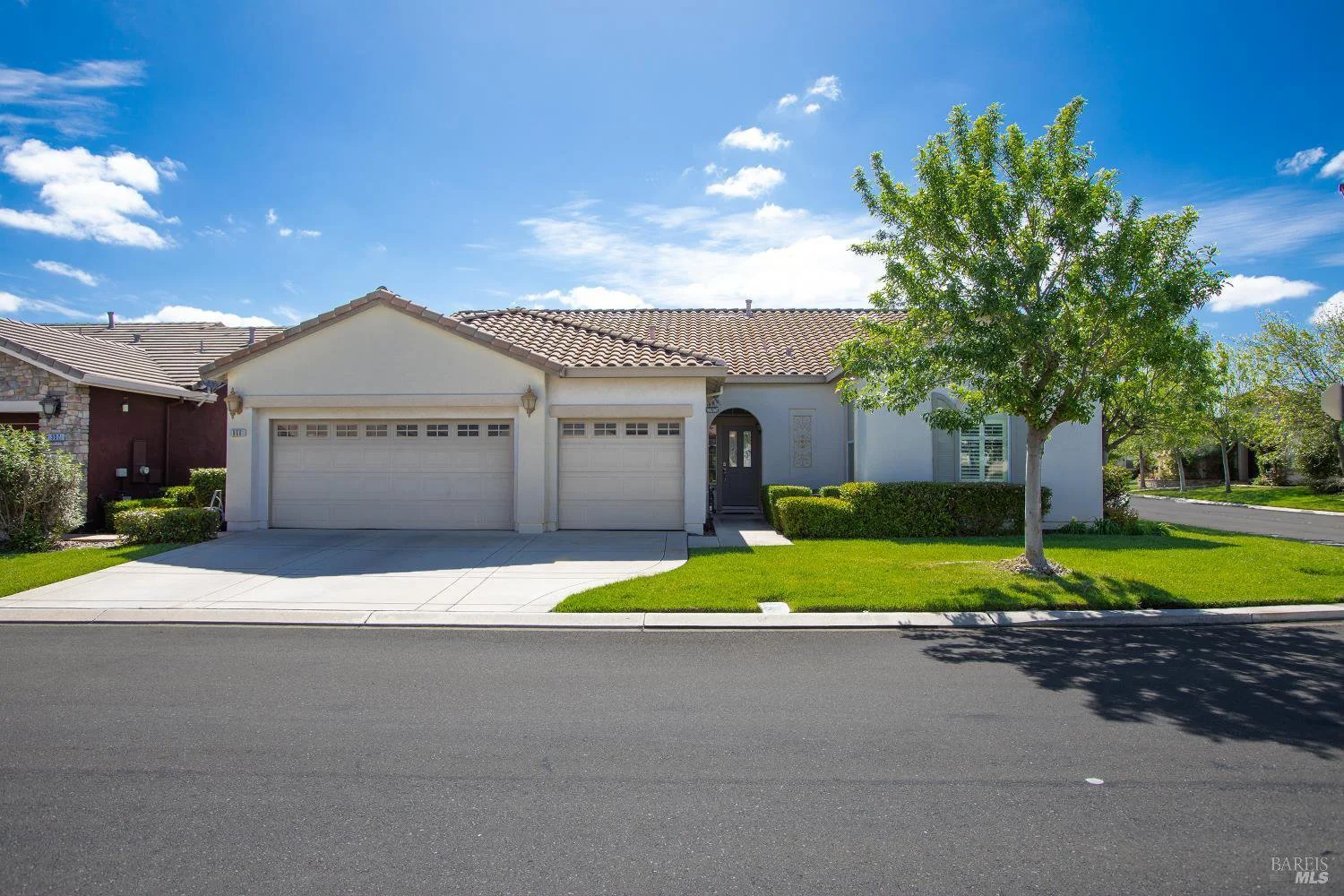 Front view of a suburban house with white exterior, two-car garage, manicured lawn, and a large tree, under a partly cloudy blue sky.