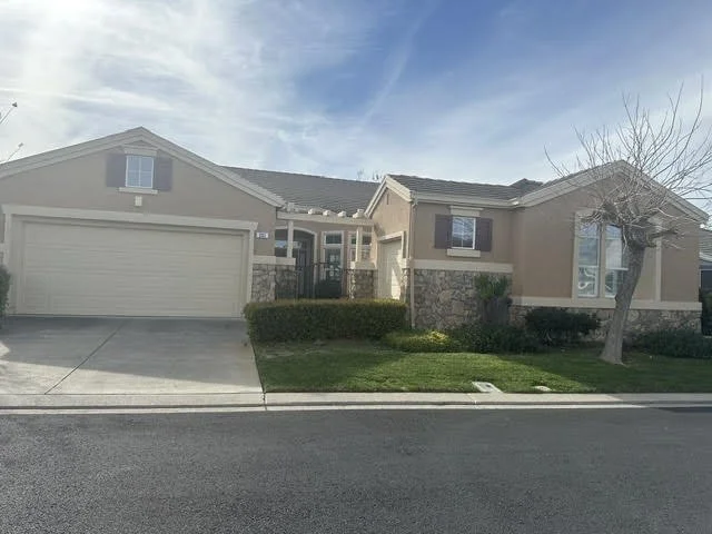 A suburban house with a two-car garage, tan exterior with stone accents, a front lawn with small bushes, and a leafless tree, under a partly cloudy sky.