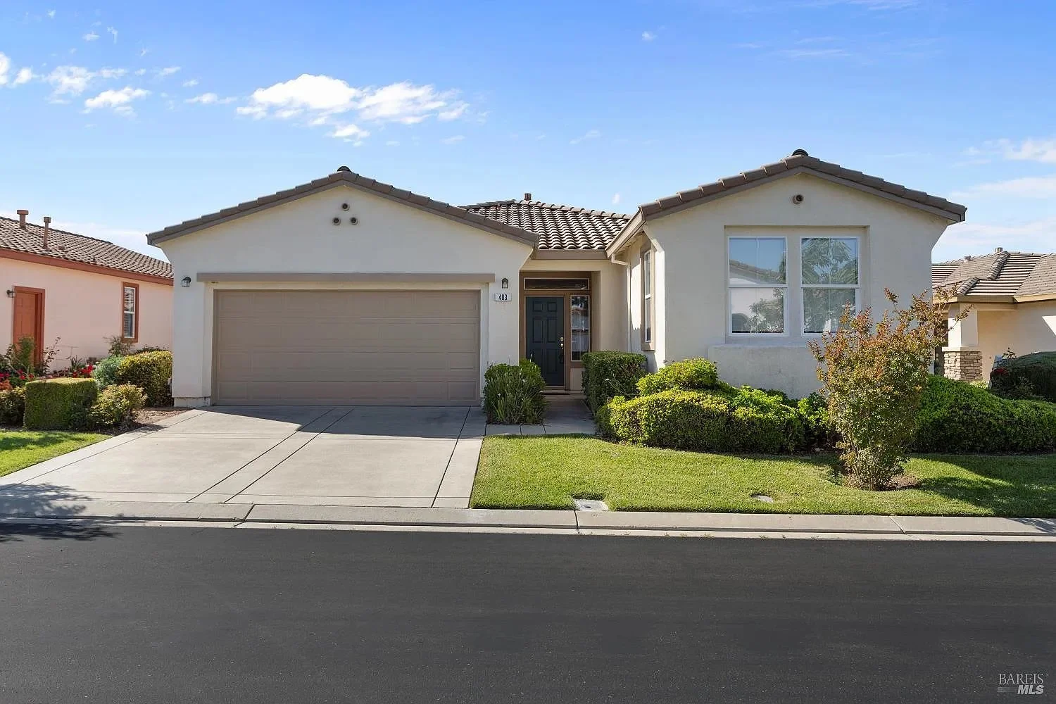 A single-story suburban house with a beige two-car garage, dark front door, and white exterior walls. The house has a well-manicured front yard with green grass, bushes, and small plants. The background shows a clear blue sky with a few clouds.