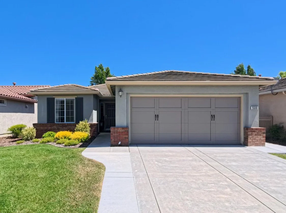Front view of a modern, white, single-family house with a two-car garage, landscaped yard, and trees, under a clear blue sky.