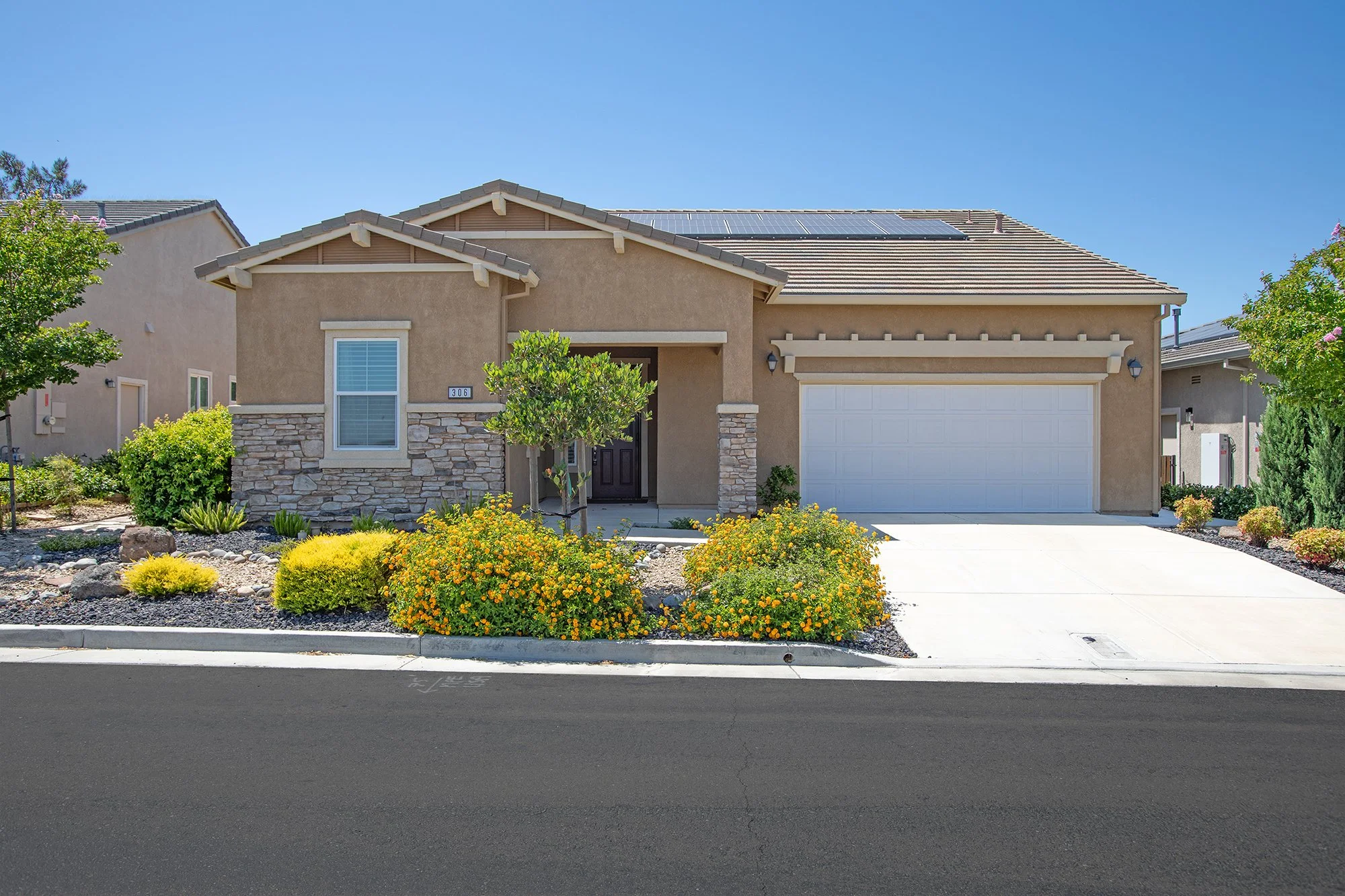 Front view of a white single-family house with a brown tiled roof, brown shutters, and attached two-car garage on a sunny day with clear blue sky, green trees, and landscaped lawn.