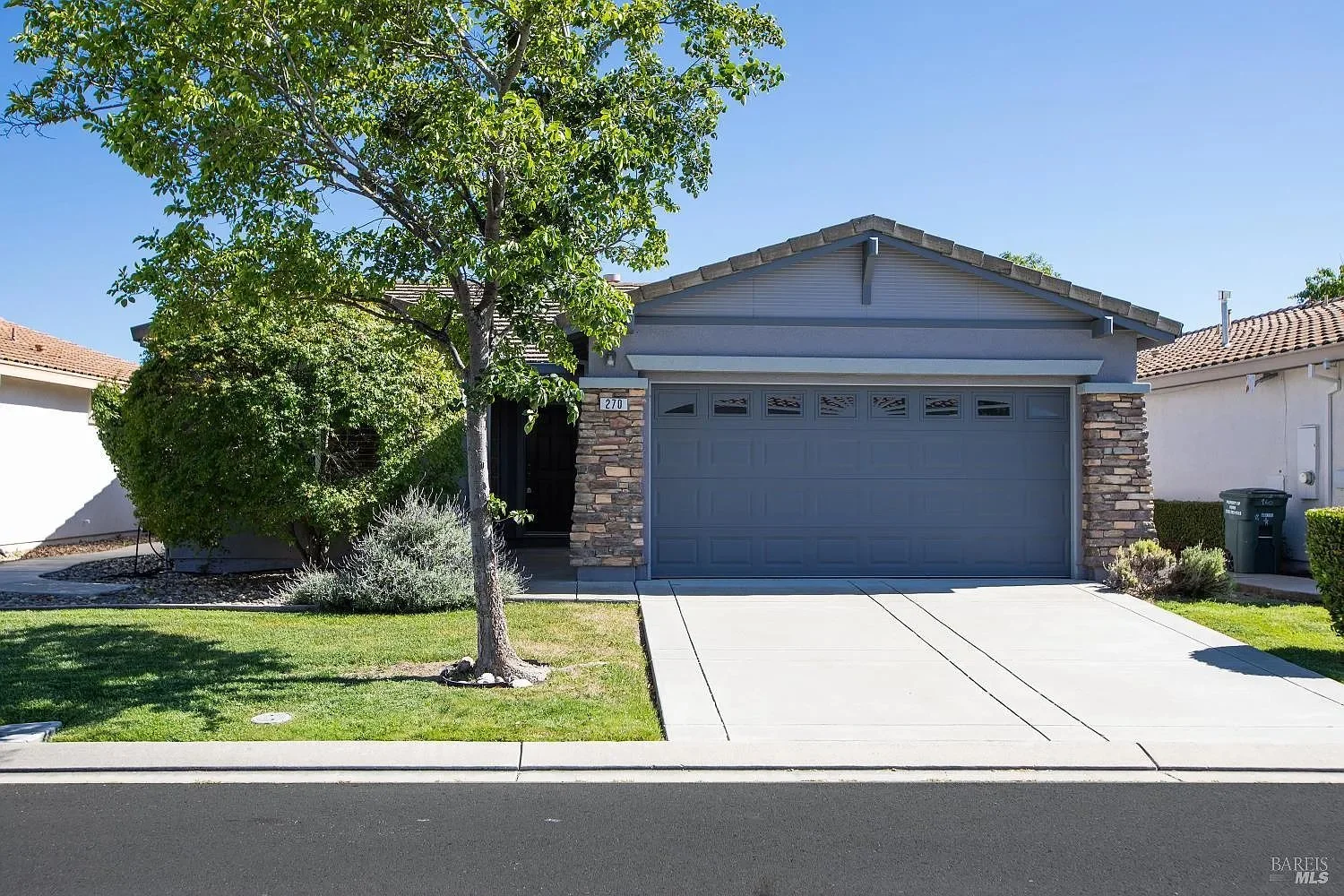 Front view of a single-story house with a gray garage door, stone accents on the columns, a small front lawn with a tree and bushes, and a concrete driveway on a clear sunny day.