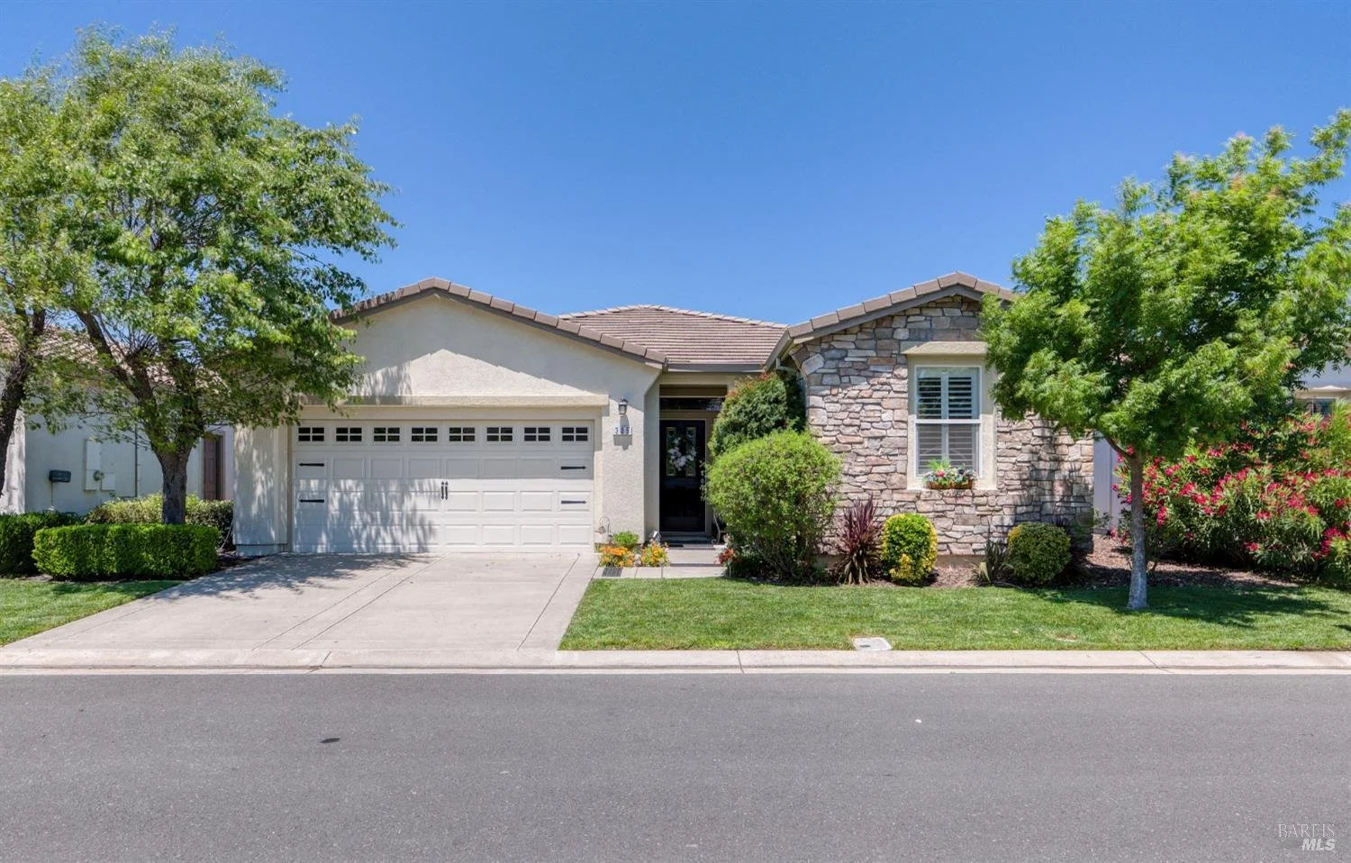 Front view of a suburban house with a two-car garage, stone and stucco exterior, surrounded by green trees and shrubs, under a clear blue sky.