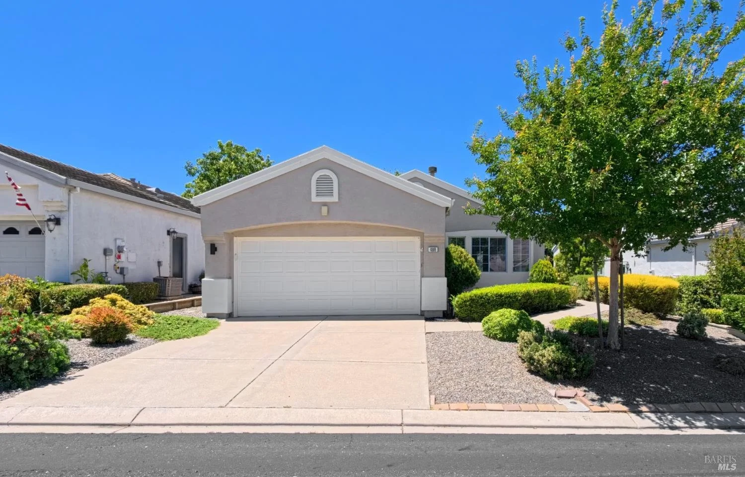 A suburban house with a beige garage door, surrounded by green trees and bushes, with a concrete driveway and a front lawn under a partly cloudy sky.