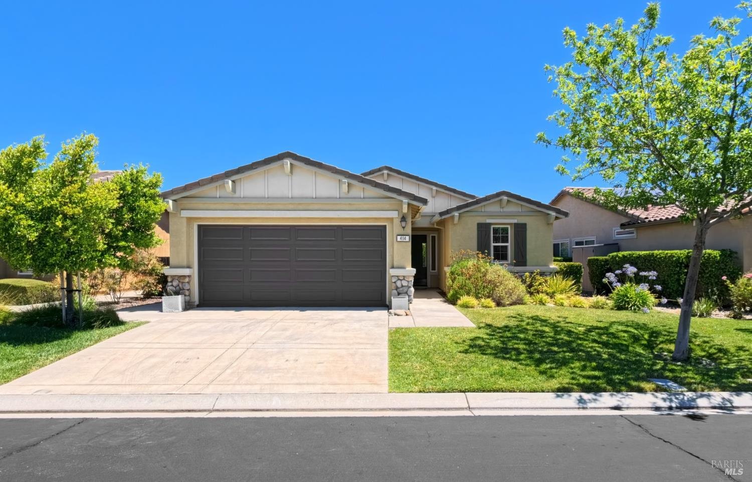 A single-story house with beige stucco exterior, a two-car garage, surrounded by a well-maintained lawn with shrubs and trees, under a clear blue sky.
