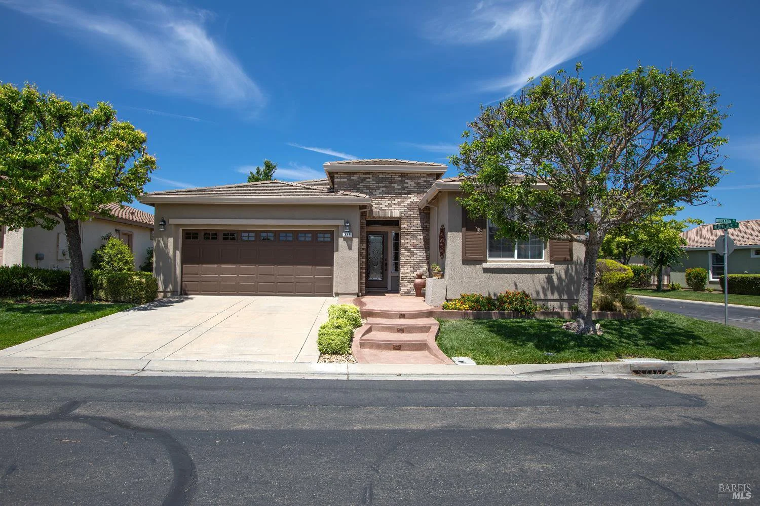 Single-story suburban house with a two-car garage, concrete driveway, front steps, and a small landscaped yard with trees and bushes under a bright blue sky.