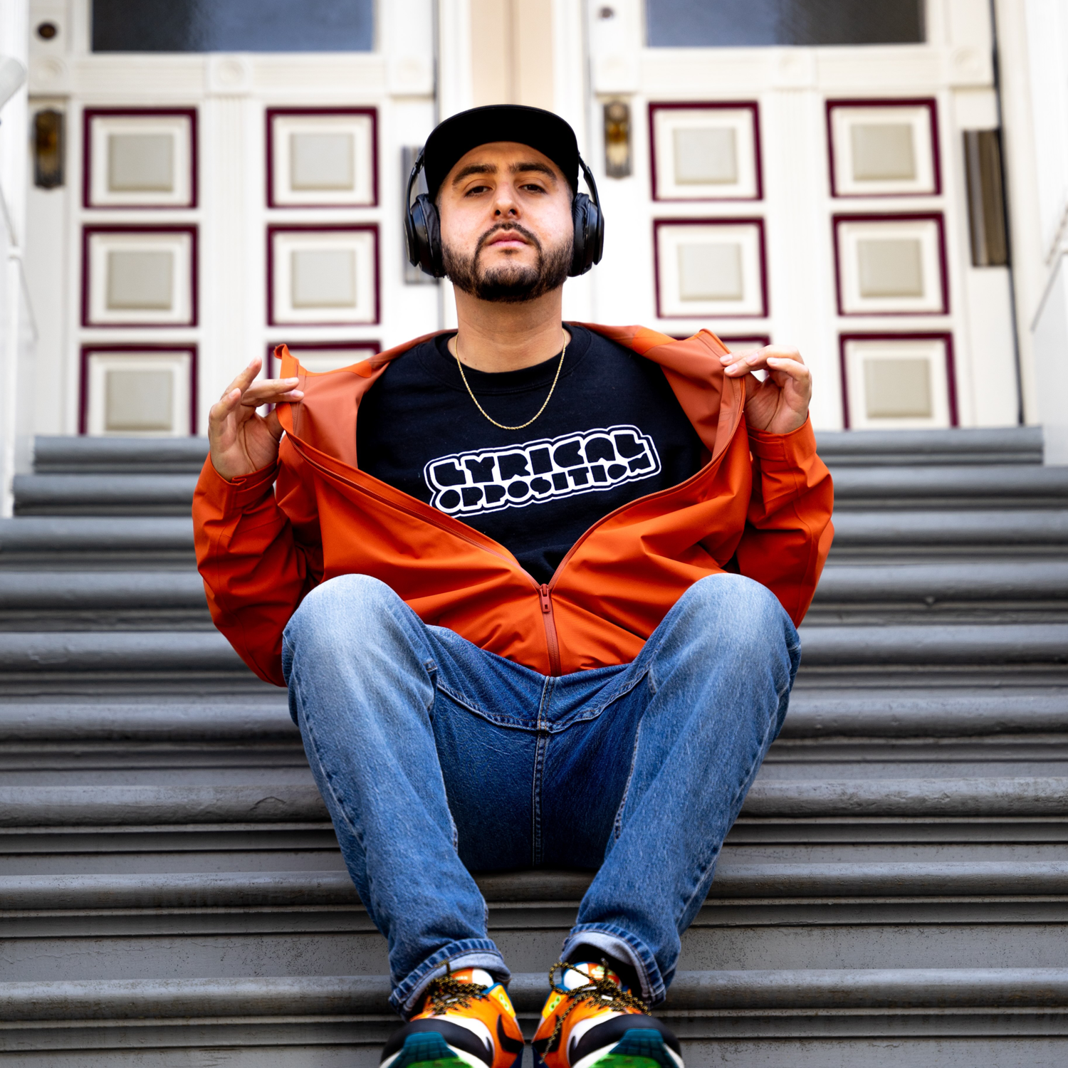 A man sitting on outdoor stairs, wearing a black t-shirt with 'Cyrical Obsess' written on it, a black cap, orange jacket open, blue jeans, colorful sneakers, and black headphones, posing in front of a decorative door.