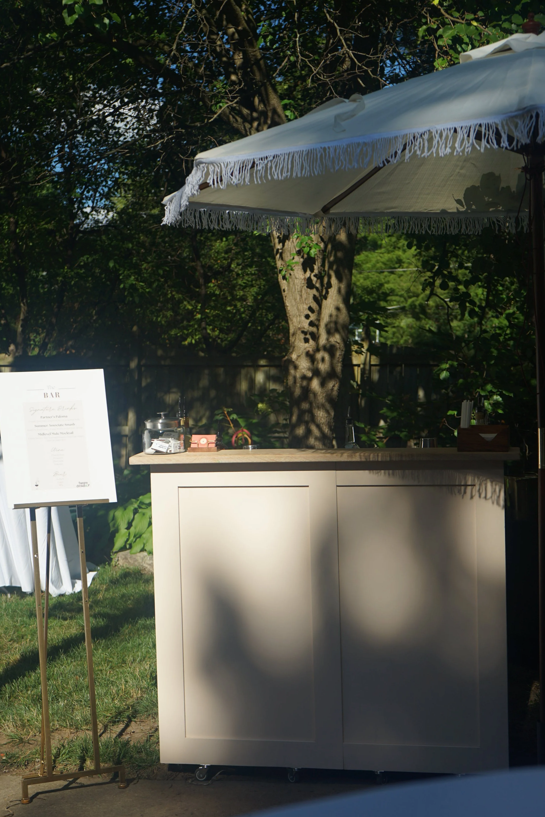 A soft pink portable bar on wheels in front of a tree and greenery next to an easel holding a poster board signature menu.