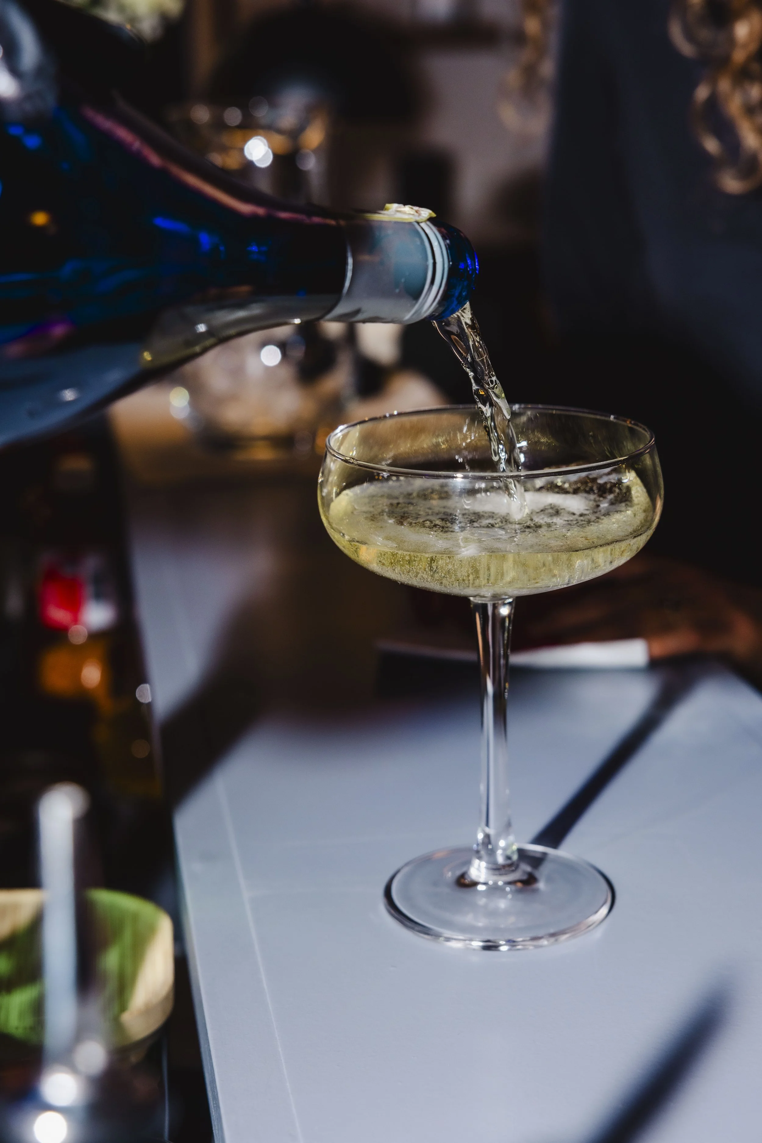 A bartender pours a glass of white bubbly wine in a coup glass as it sits atop a gray bar top.