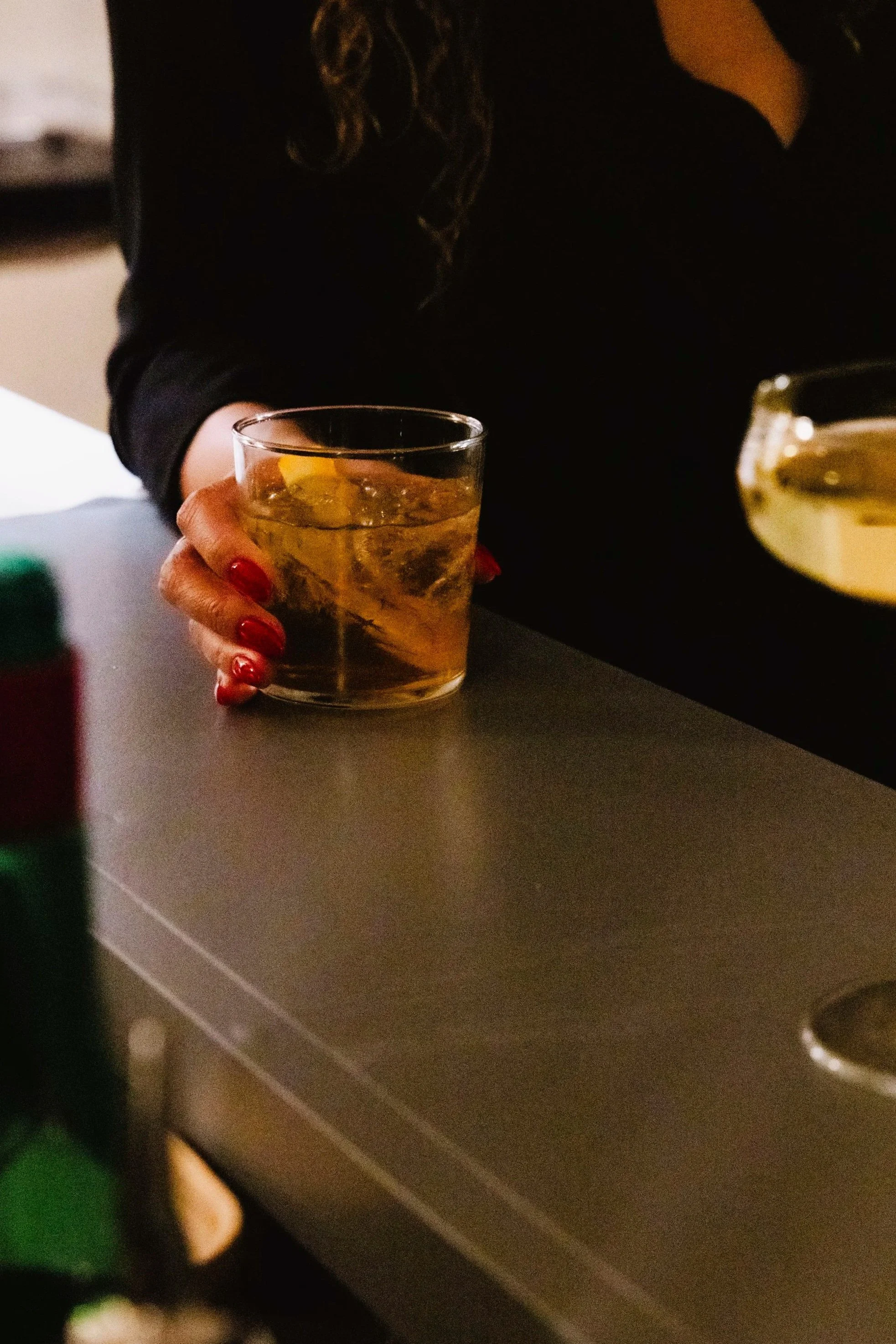 A glass of sparkling water with a lemon slice resting on top, placed on a light-colored surface. Partially visible bowls of rice and noodle salad are seen on the left side.
