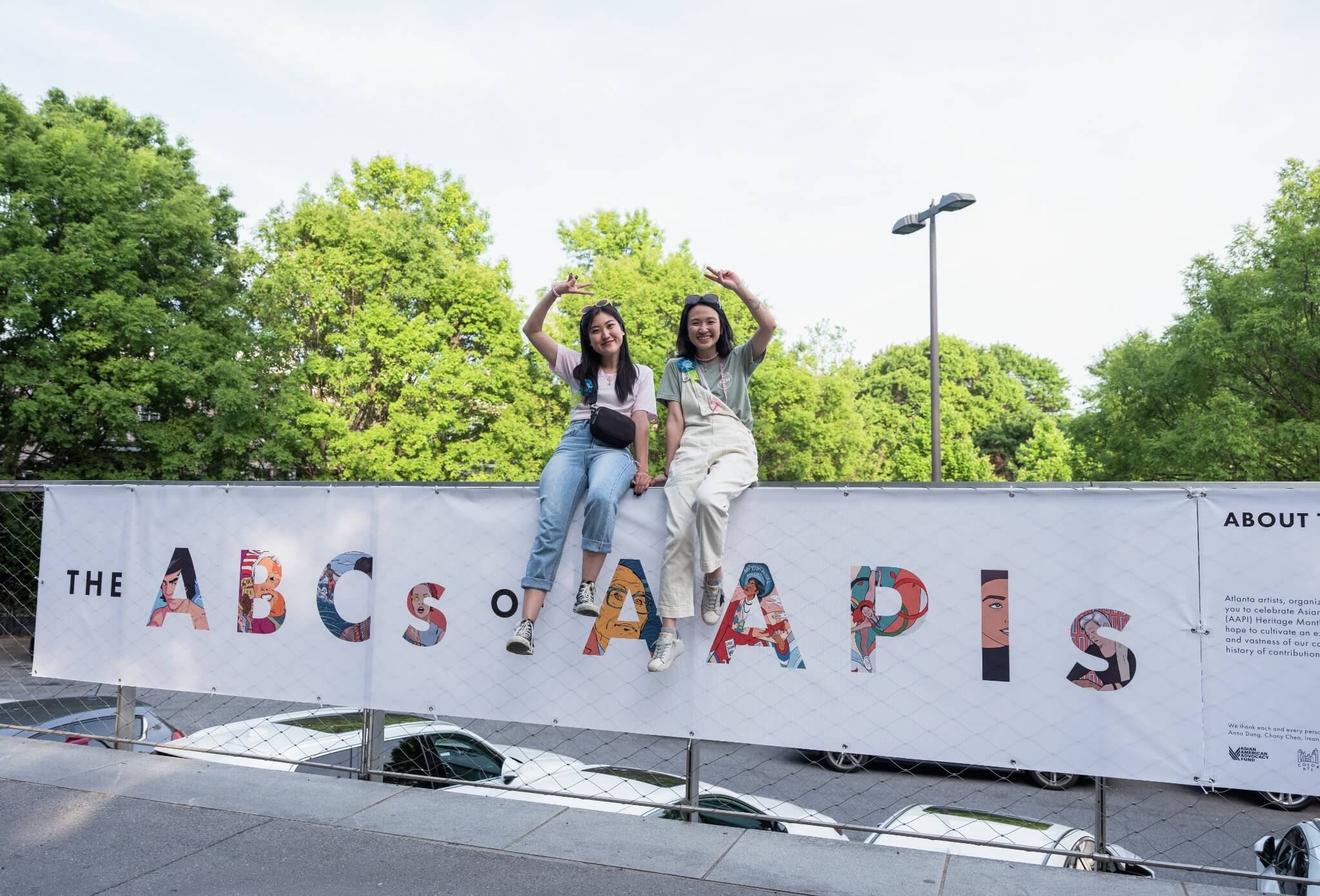  Co-creative directors Arah Kang and Kavi Vu sit atop the BeltLine fence where a large banner hangs with the words “The ABCs of AAPIs”&nbsp; 