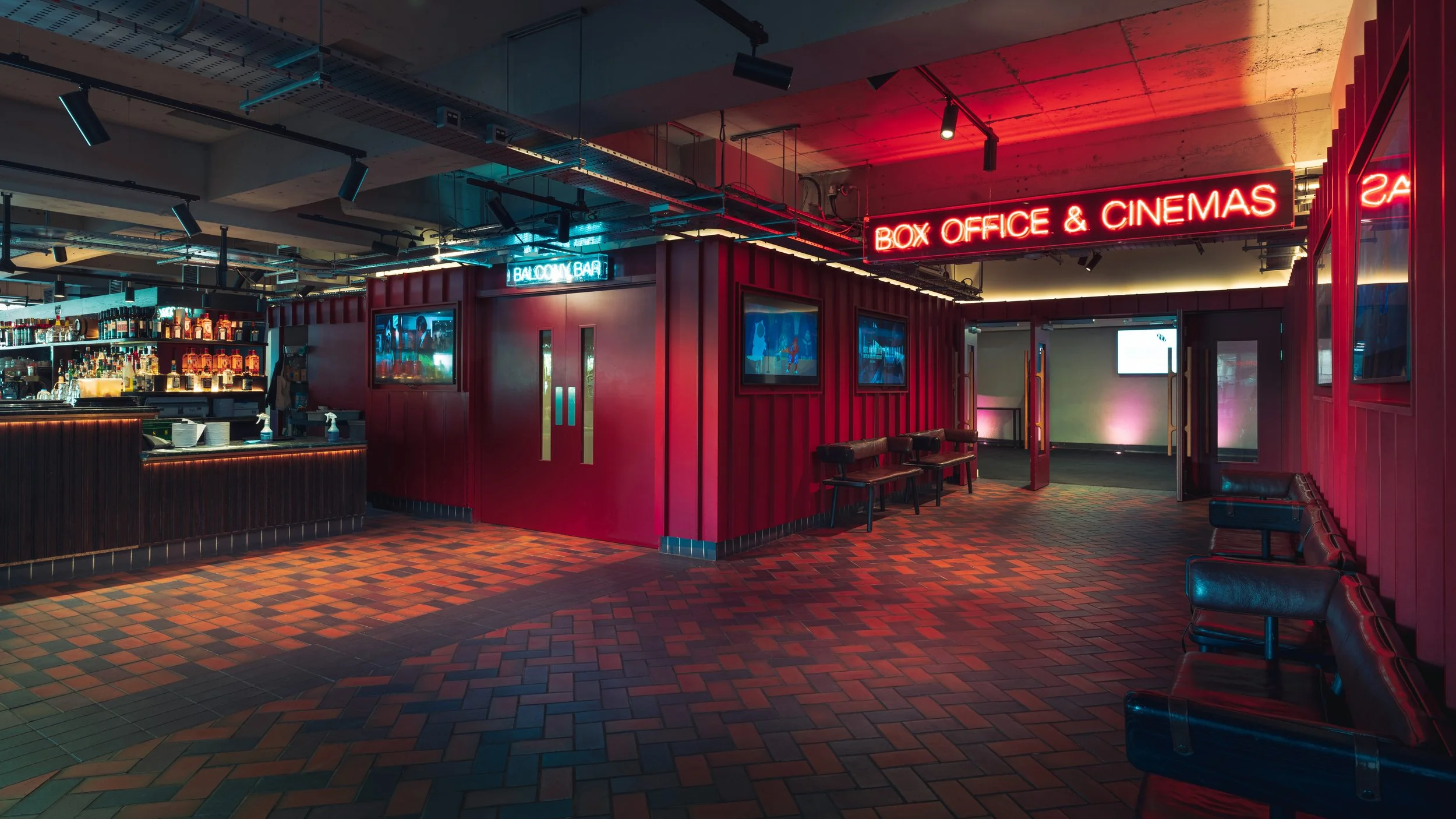 Interior of a dimly lit entertainment venue with a bar area on the left, red walls, multiple television screens, and neon signs that say 'BOX OFFICE & CINEMAS' and 'BALCONY & BAR'.