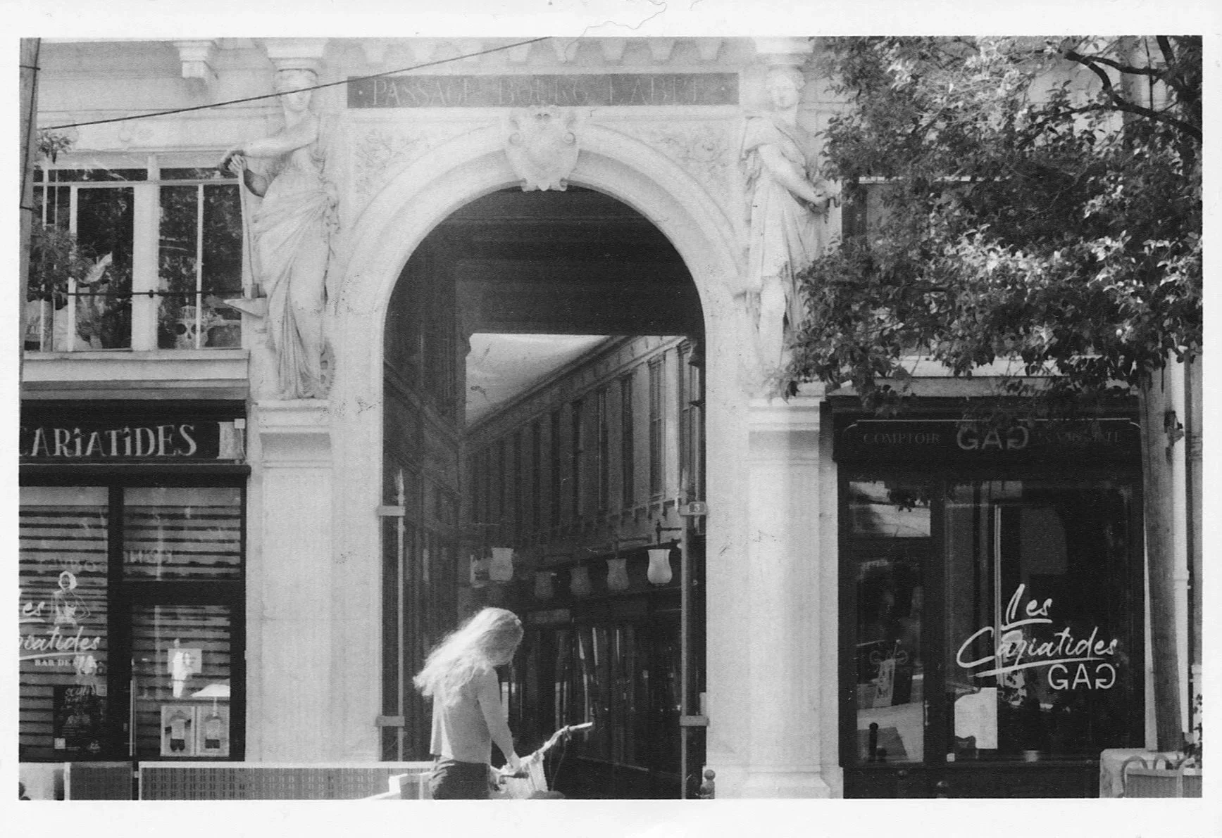 Black and white photograph of an ornate Parisian archway with shops either side. Two statues of women hold up the facade of the arch. A young woman stands in the centre foreground, facing away.