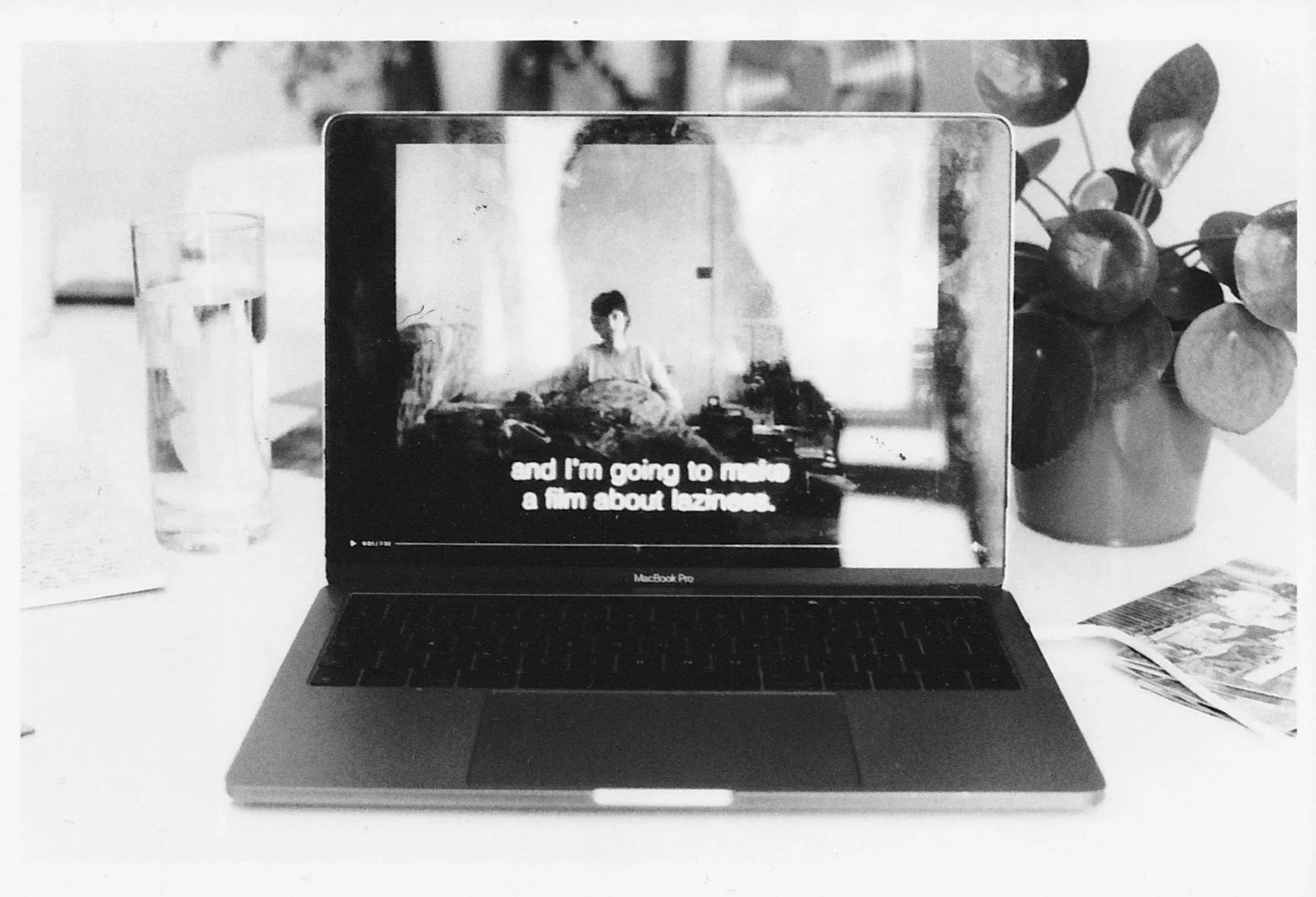 Black and white photo of the same previous scene. The laptop now shows another moment from the same film, Portrait of a Lazy Woman by Chantal Akerman. On screen the woman is lying in bed, subtitles read: “and I’m going to make a film about laziness”