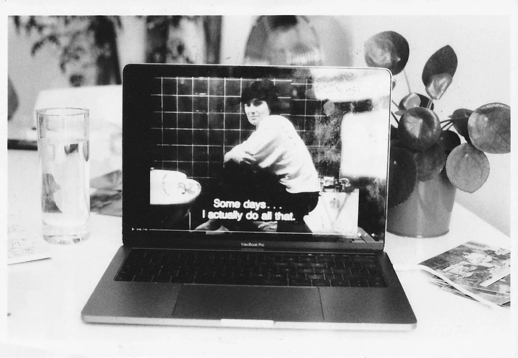 Black and white photo of a laptop. On screen there is a woman crouching in a tiled room, subtitles read “Some days…I actually do all that.” Next to the laptop is a full glass of water and a small potted plant.