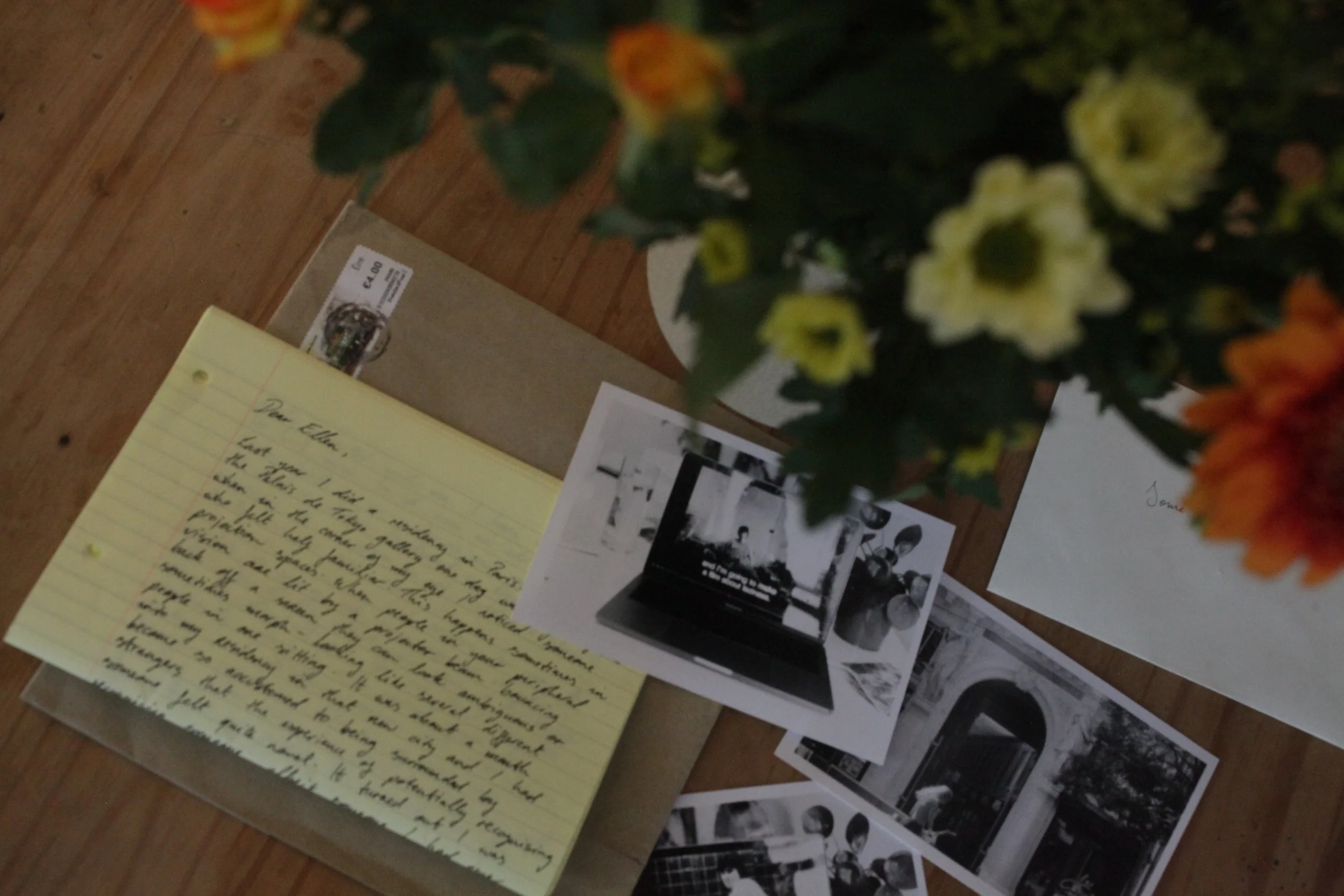 Overhead photograph of an envelope, handwritten letter, and overlapping black and white photographs on a wooden table. In the upper right of the image a vase of yellow and orange flowers partially obscures the table.