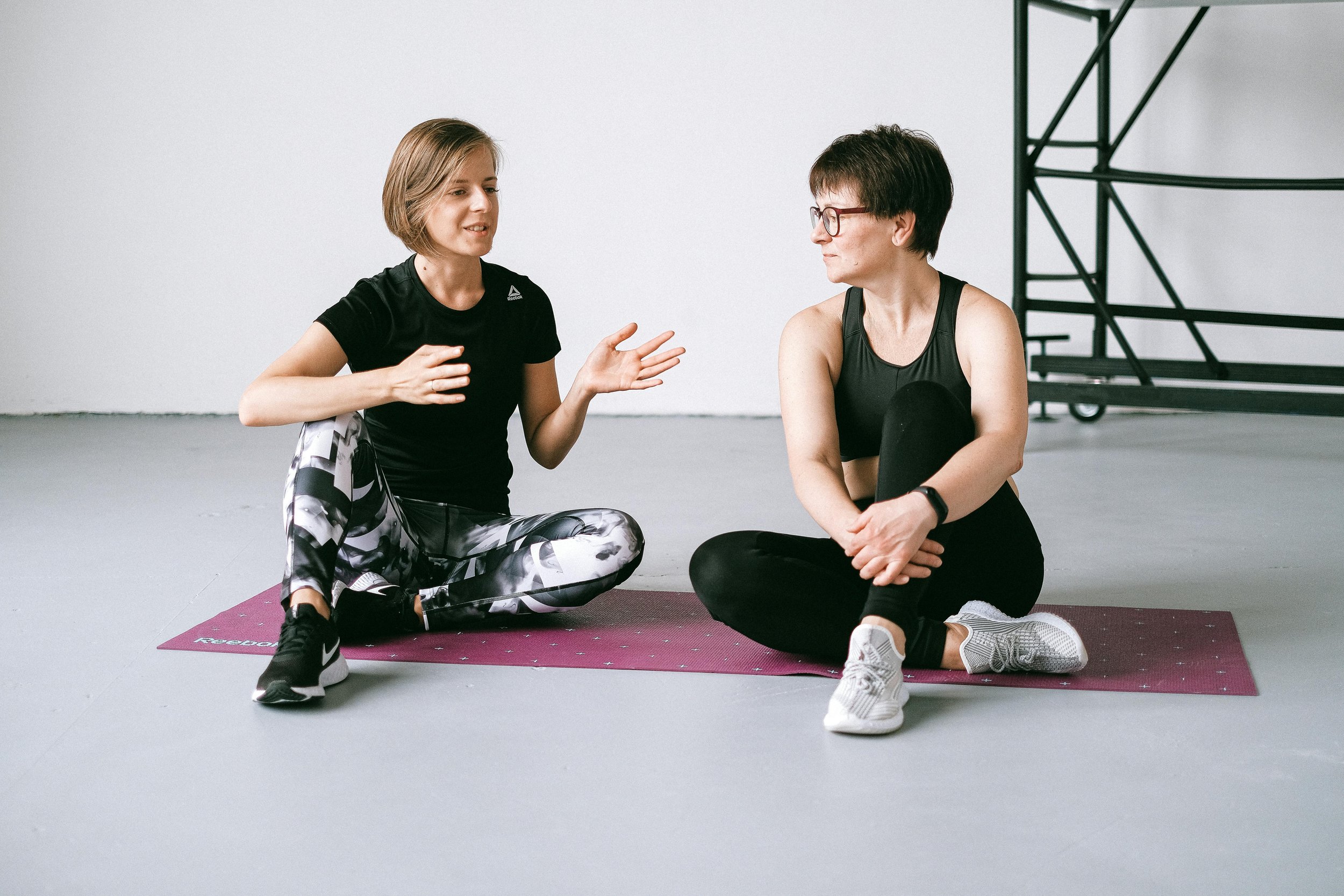 Two women sitting on yoga mats in a gym, having a conversation. One woman wears athletic clothes with leggings and sneakers, the other wears a tank top and glasses.