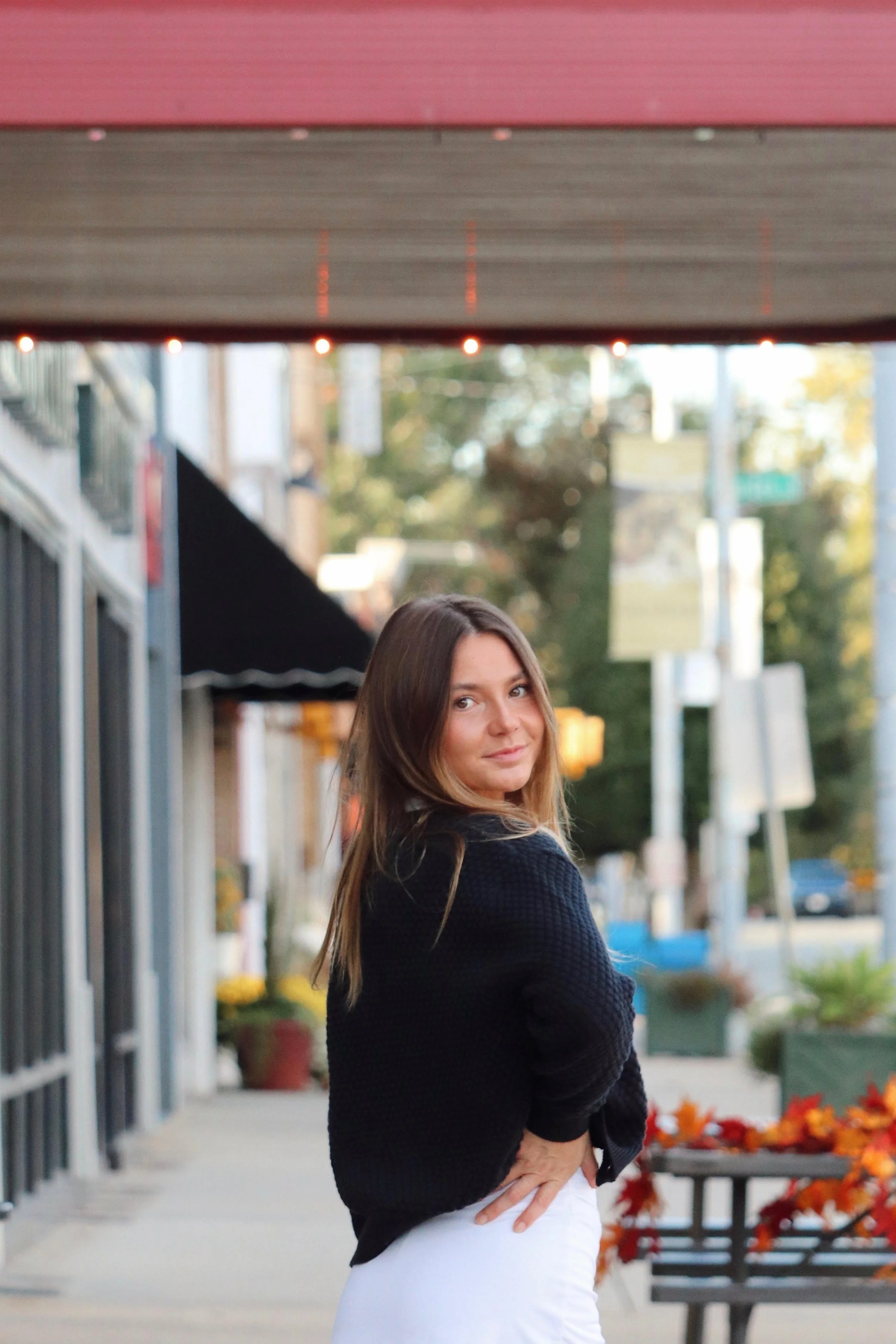A woman with long brown hair poses outdoors in a black sweater and white pants, holding her hands on her hips, with a city street, trees, and fall foliage in the background.