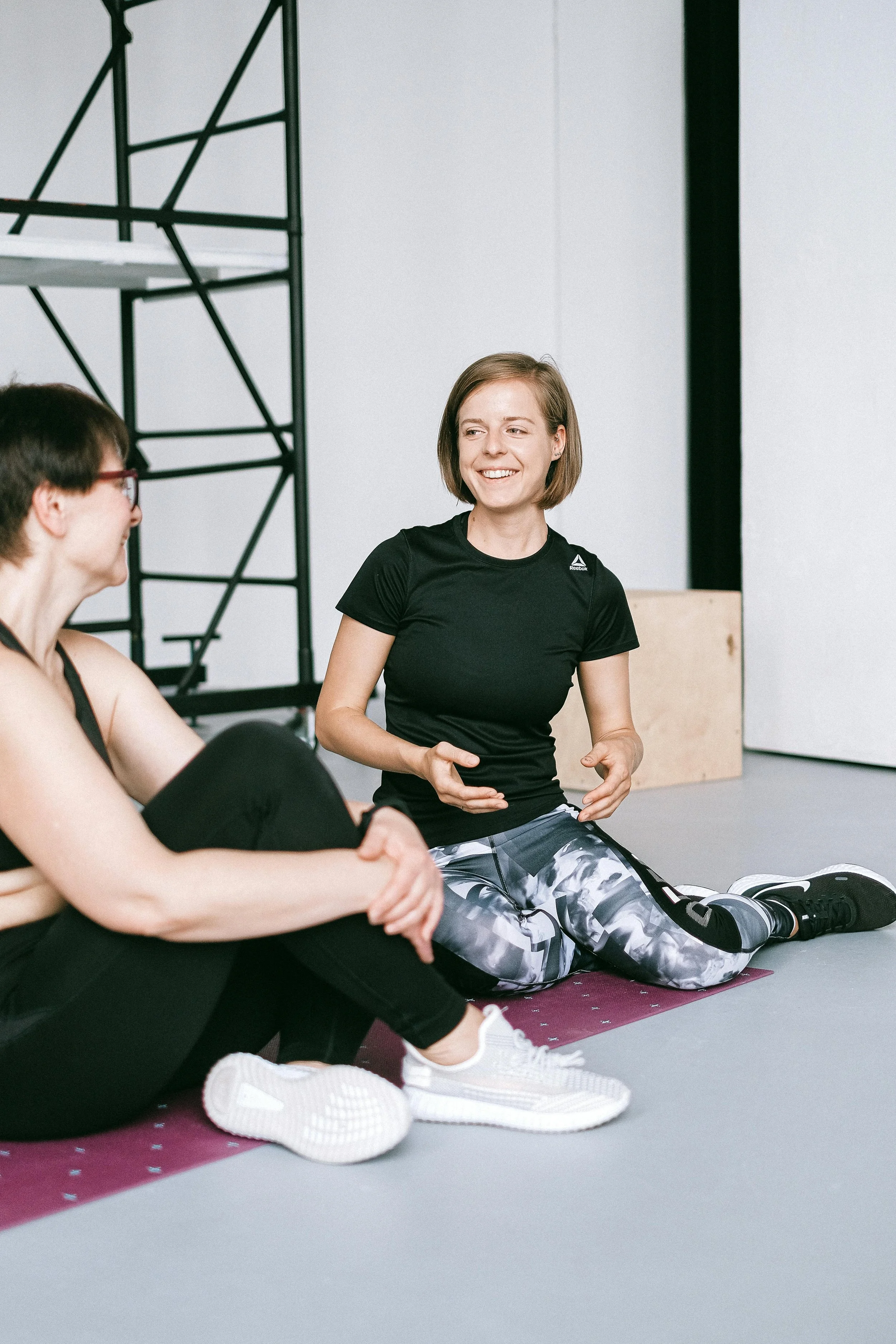 Women sitting on exercise mats in a fitness studio, engaging in a fitness class or workout session.