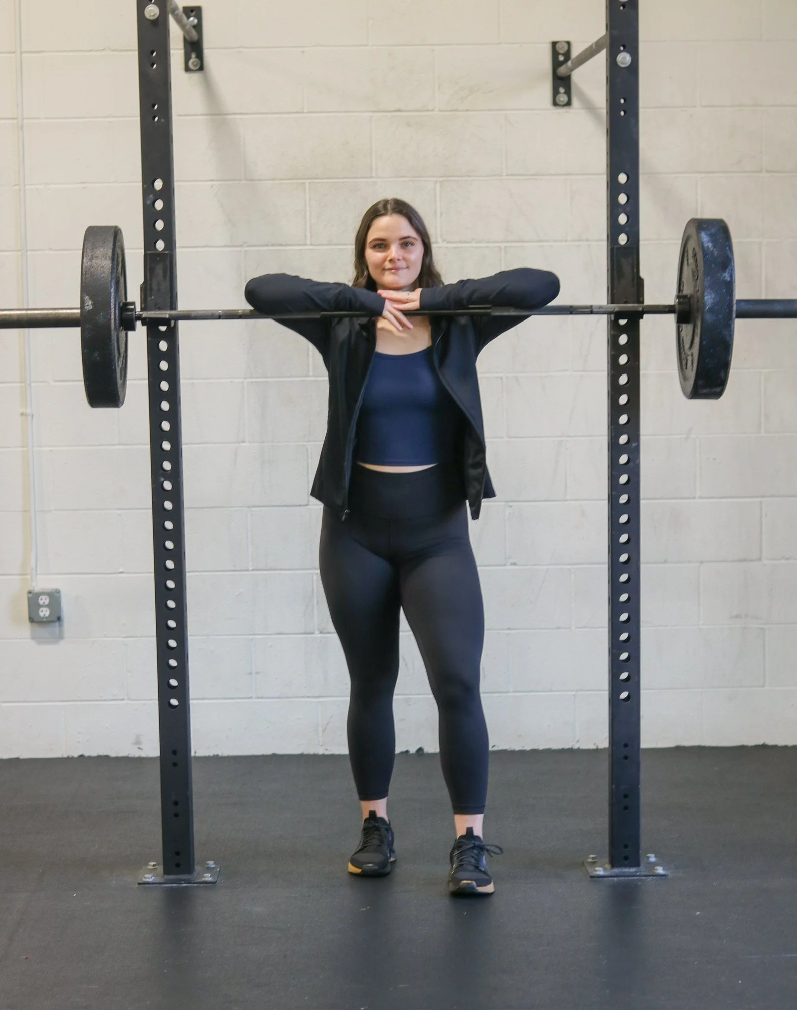 A woman standing behind a barbell with weights at a gym, with her arms resting on the bar in a stretching pose.