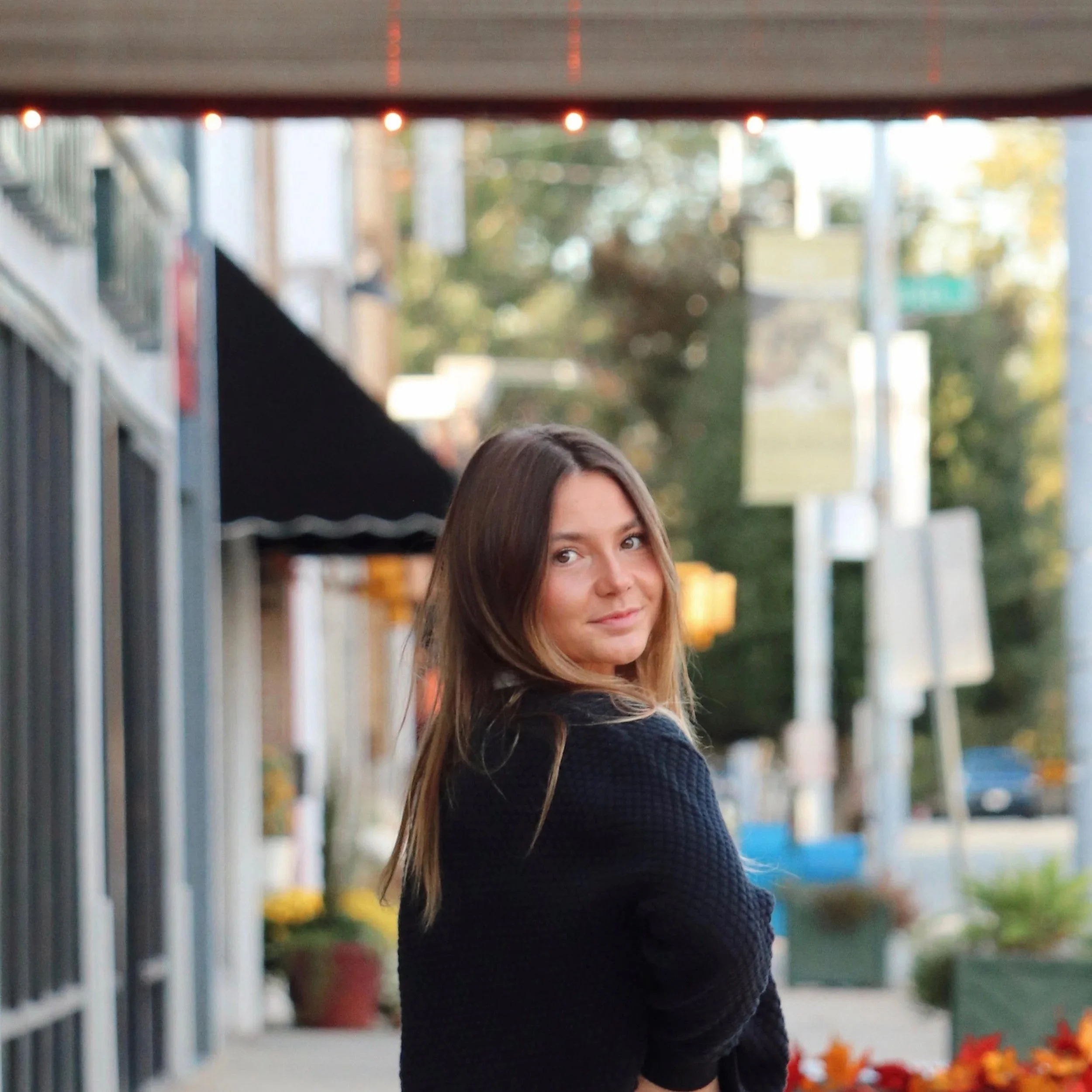 A woman with long brown hair wearing a black sweater looks over her shoulder at the camera in front of a blurred outdoor setting with trees and buildings, under a structure with small red lights.
