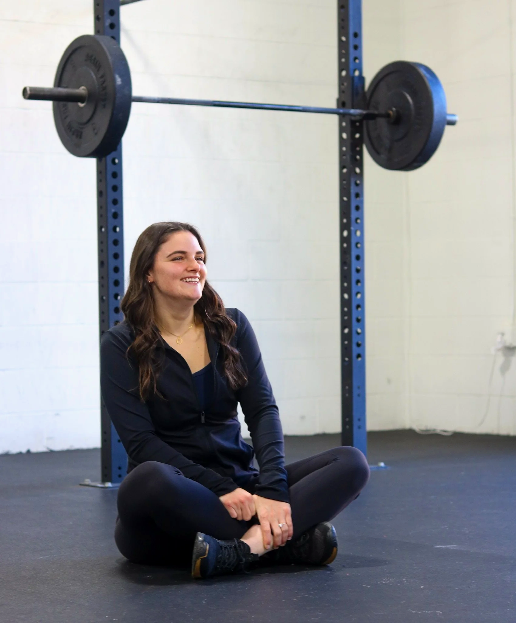 A woman sitting on the floor in a gym, holding her ankle with a pained expression, with a barbell on the rack behind her.