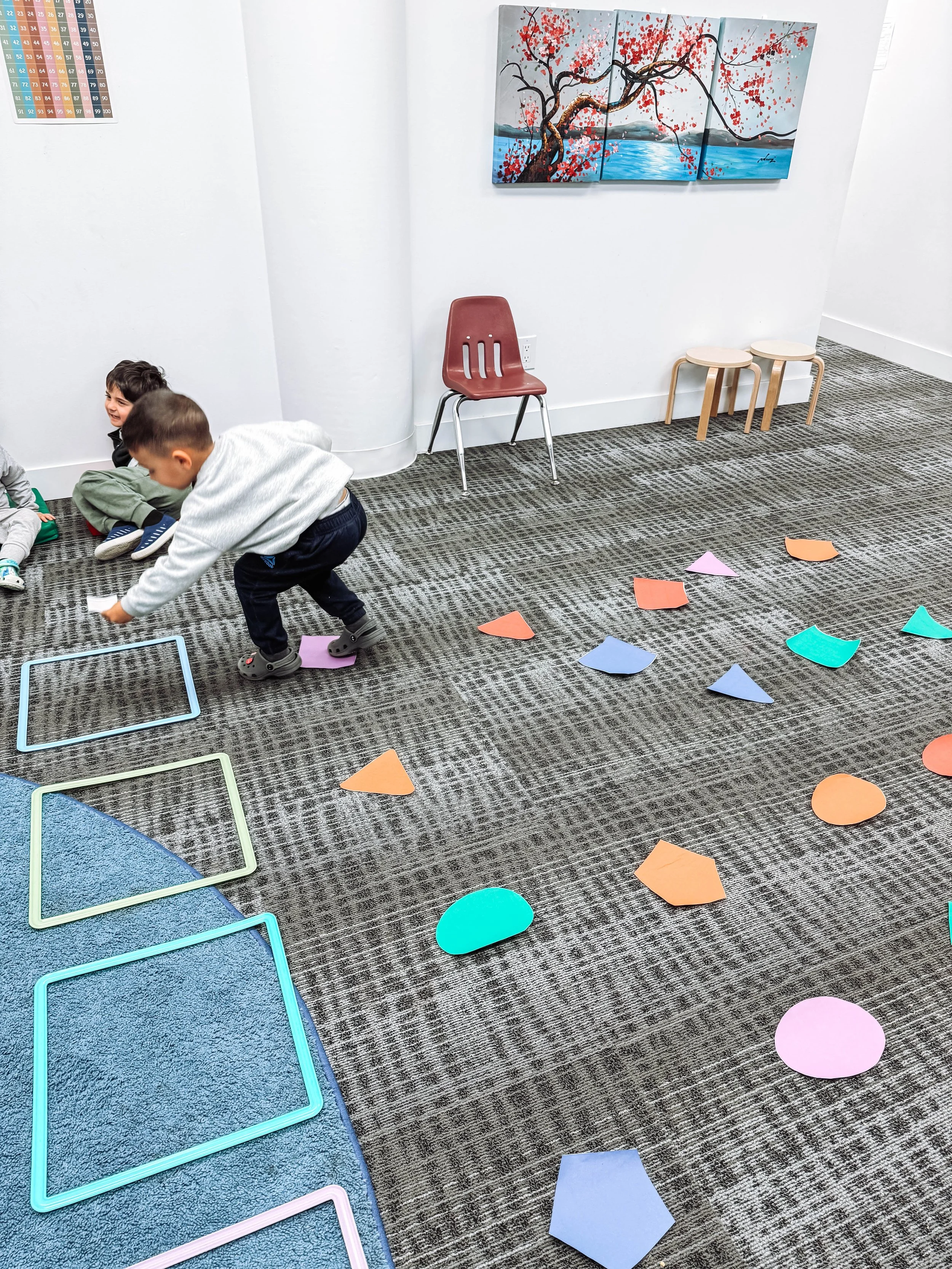Children playing with colorful shapes on a carpeted indoor floor in a classroom or playroom. There are chairs and wall art in the background.