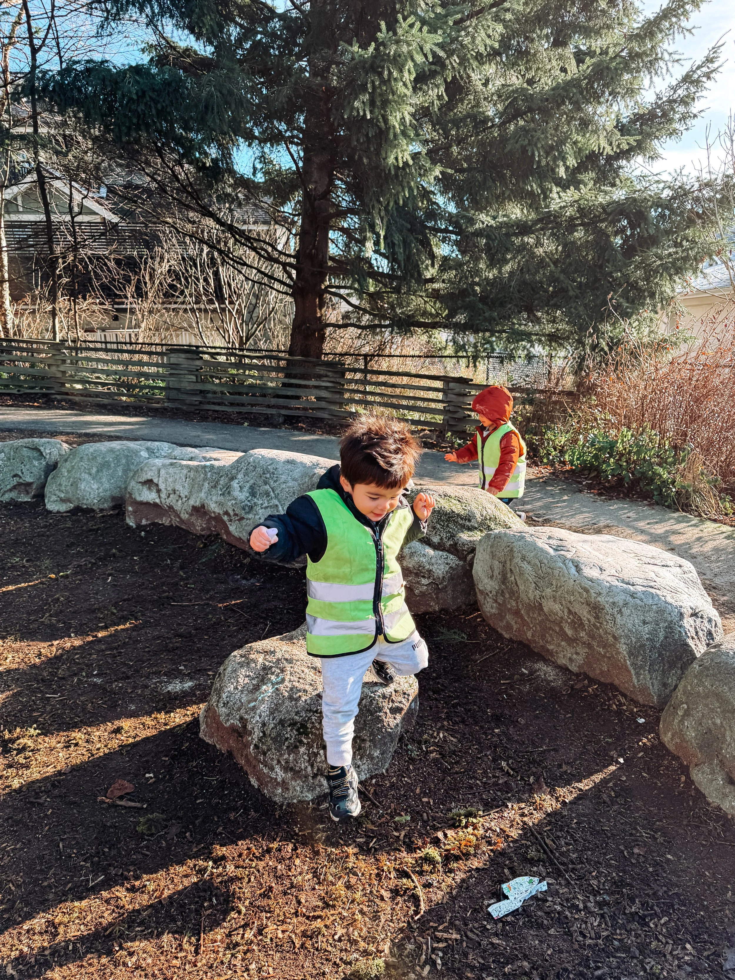 Two children playing outdoors on large rocks, wearing bright yellow and orange safety vests, with a large tree and a wooden fence in the background.