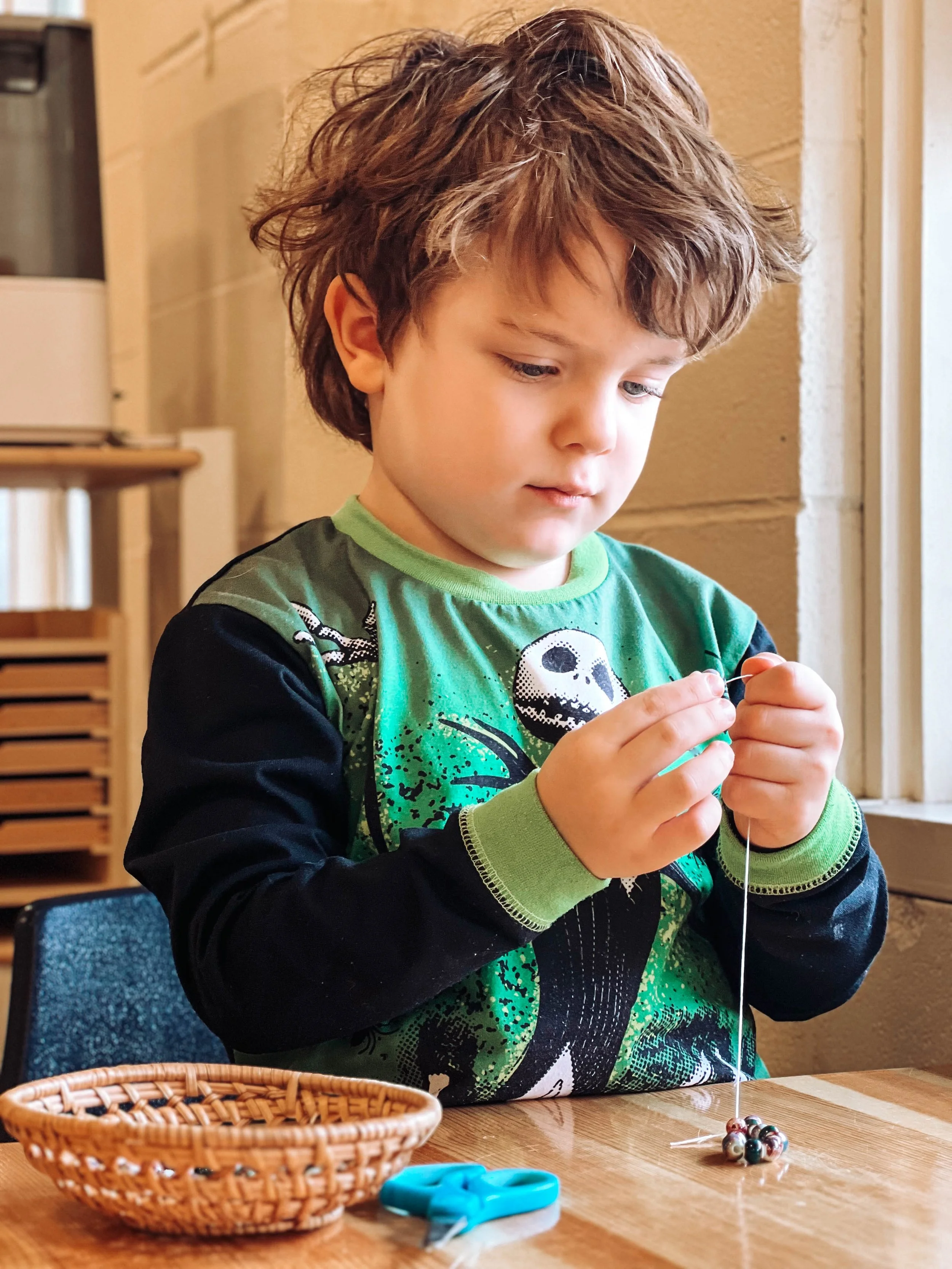 Young boy with curly hair wearing a green and black shirt, sitting at a wooden table, stringing beads into a necklace or bracelet, with a small woven basket and scissors on the table.