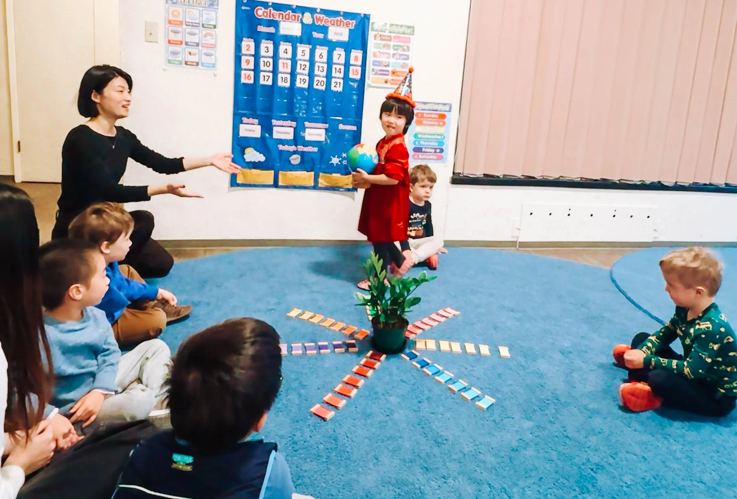 A classroom scene where a young girl in a red dress and birthday hat stands on a small wooden block, holding a globe, as a woman gestures towards her. Several children are sitting on a blue carpet, watching the girl, with some holding orange balls. B