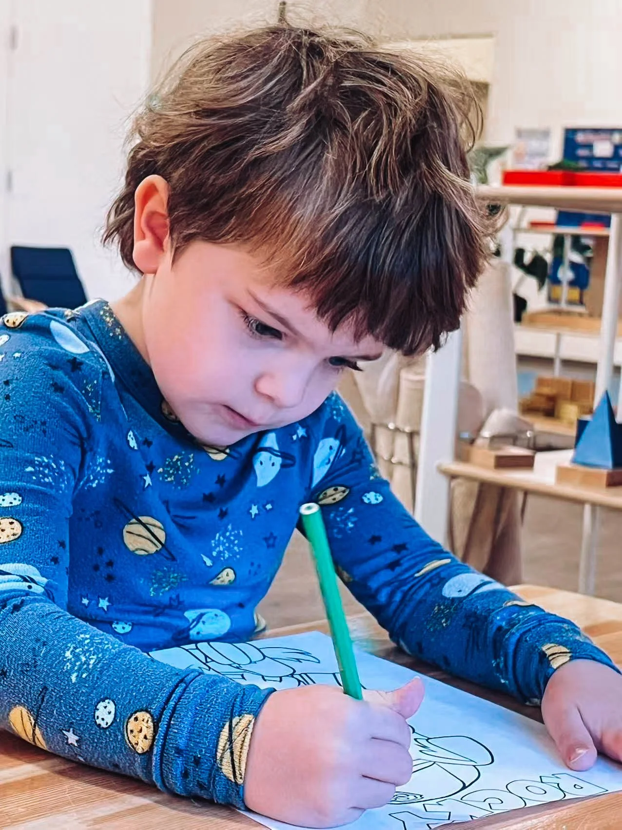 Young boy with brown hair wearing a blue space-themed shirt coloring on paper with a green crayon in a classroom or art space.