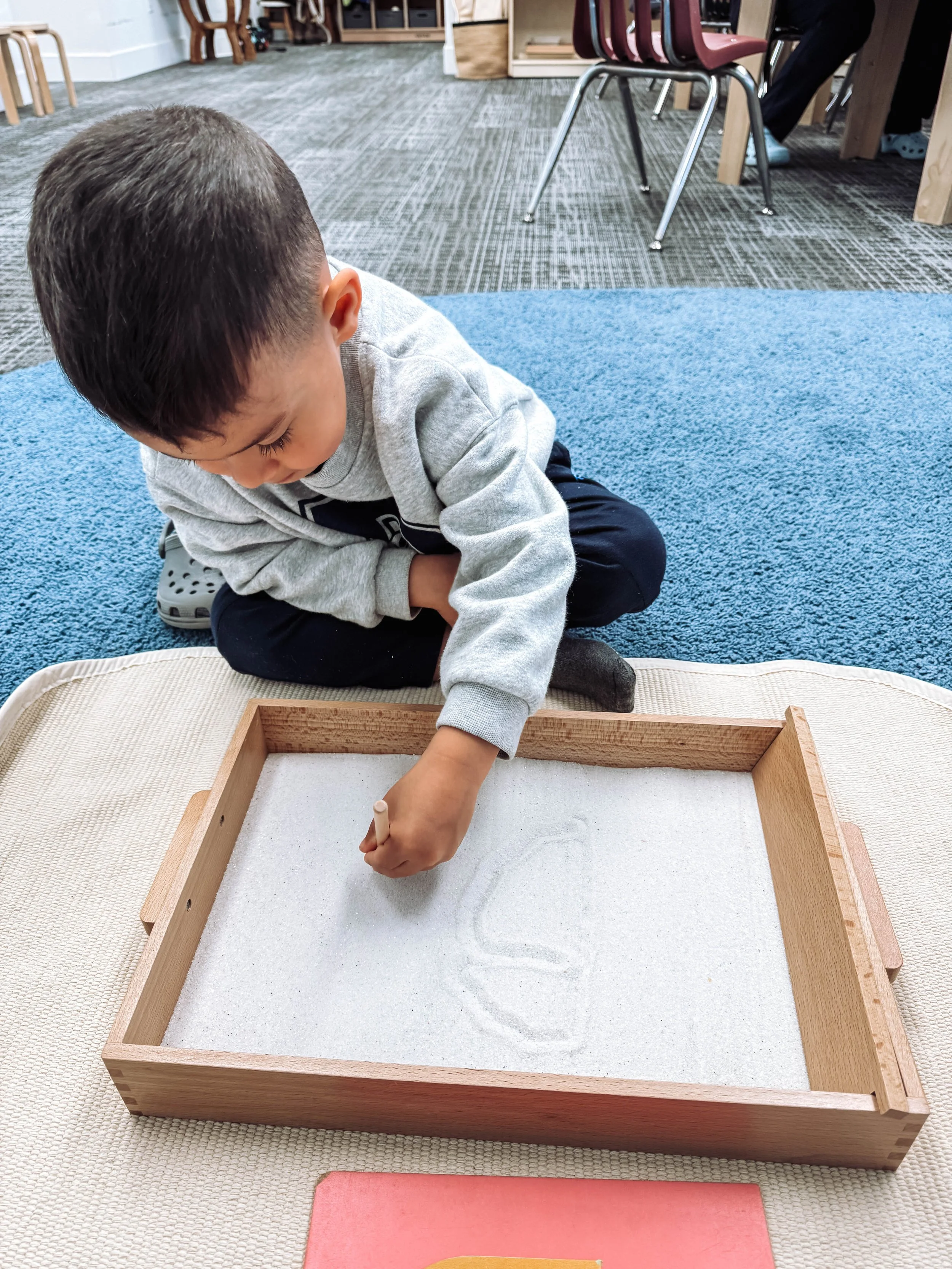 A young boy with dark hair, wearing a gray sweatshirt and black pants, sitting cross-legged on a blue carpet, drawing in a wooden sandbox with white sand using a stick.