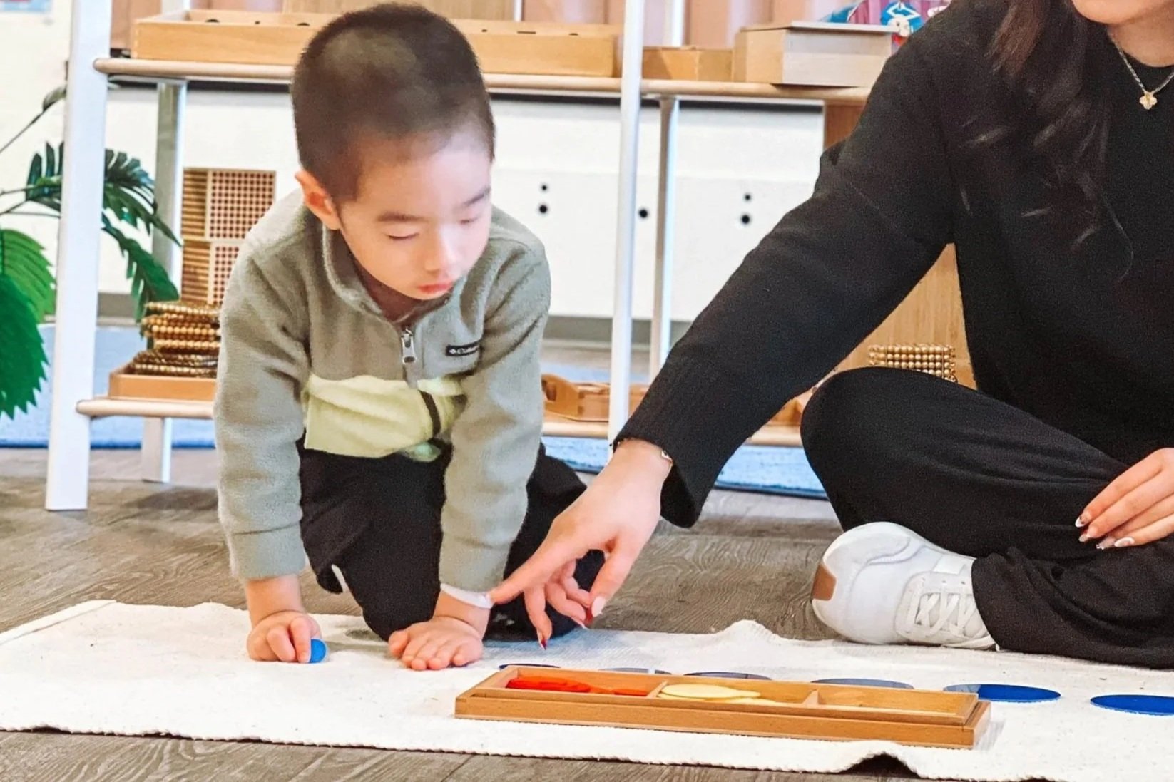 A woman and a young boy sitting on the floor in a classroom, playing with educational toys and game pieces on a beige rug.