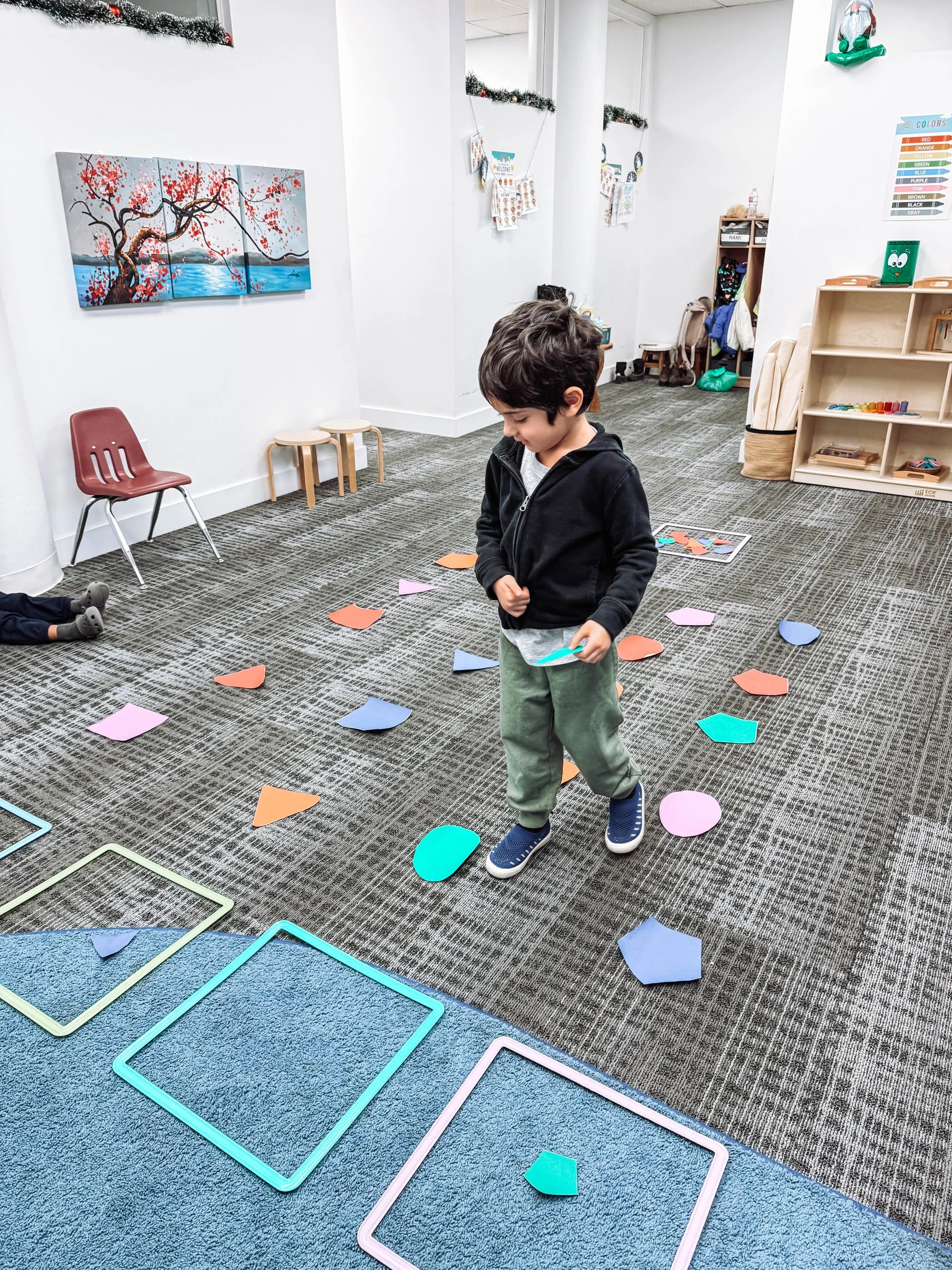 A young boy in a black hoodie and green pants is standing on a carpeted floor with colorful paper shapes scattered around him. The shapes include circles and squares in pastel colors. The room has a white wall with a framed painting of a tree with re