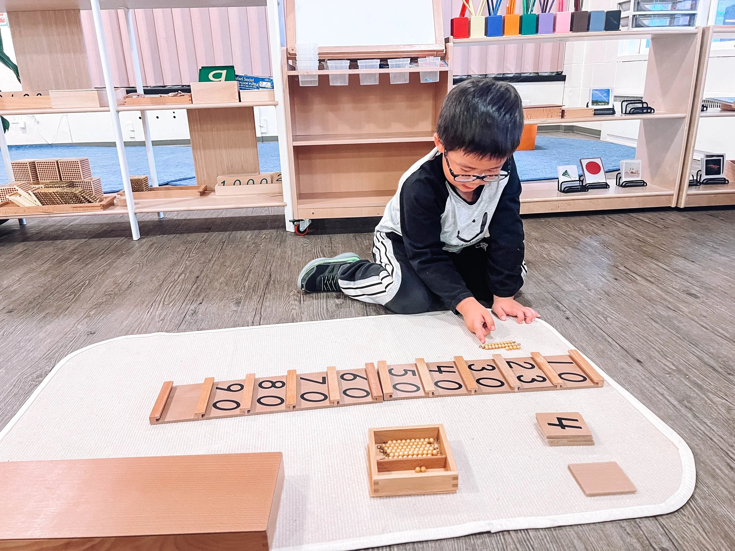 A young boy kneels on a white rug, using educational toys that include numbered tiles and a bead abacus, in a classroom with wooden shelves and educational materials in the background.