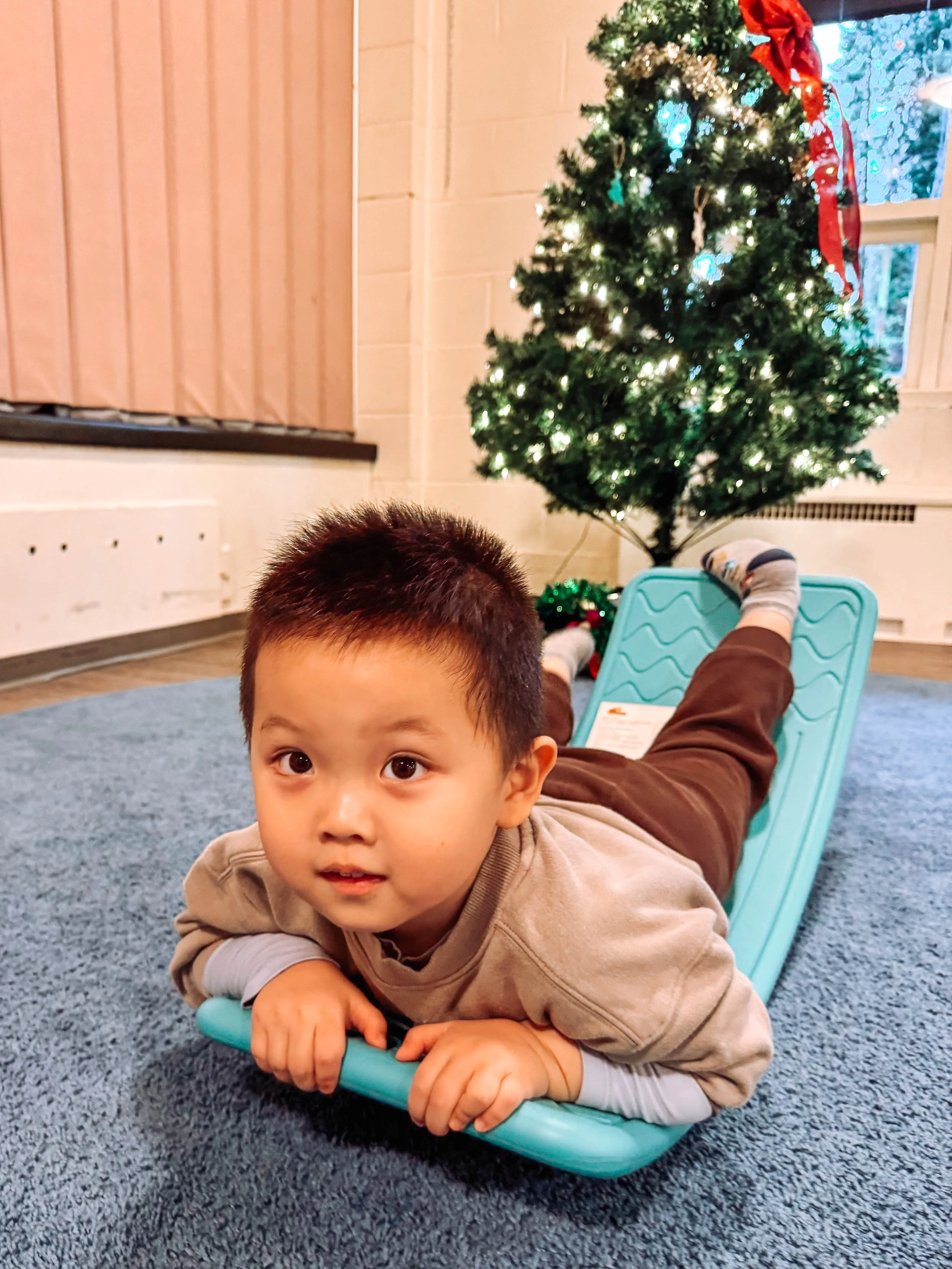 Child lying on a blue padded slide, looking at the camera, with a decorated Christmas tree in the background.