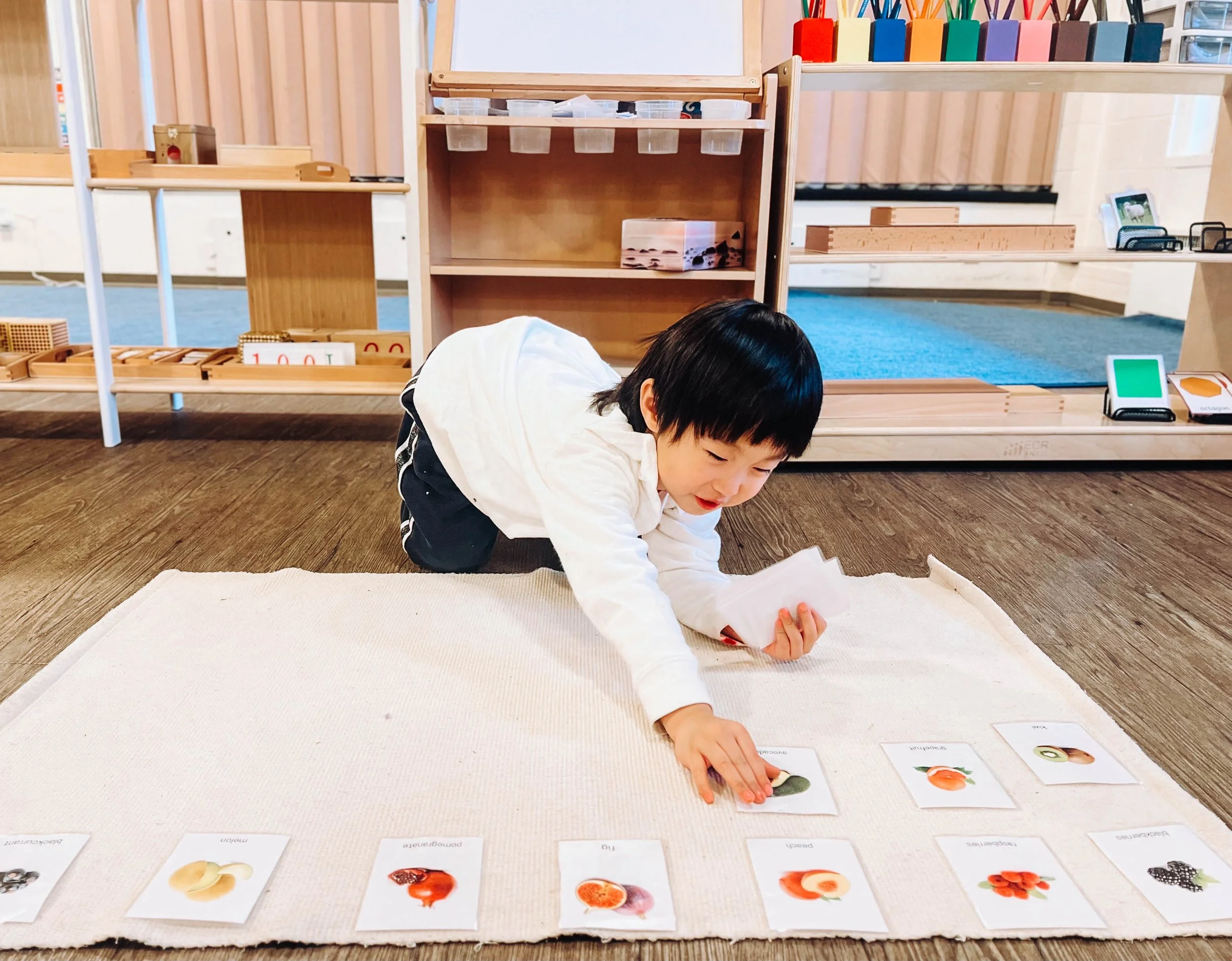 Child crawling on a white rug, playing with picture cards of fruits and vegetables in a classroom setting.
