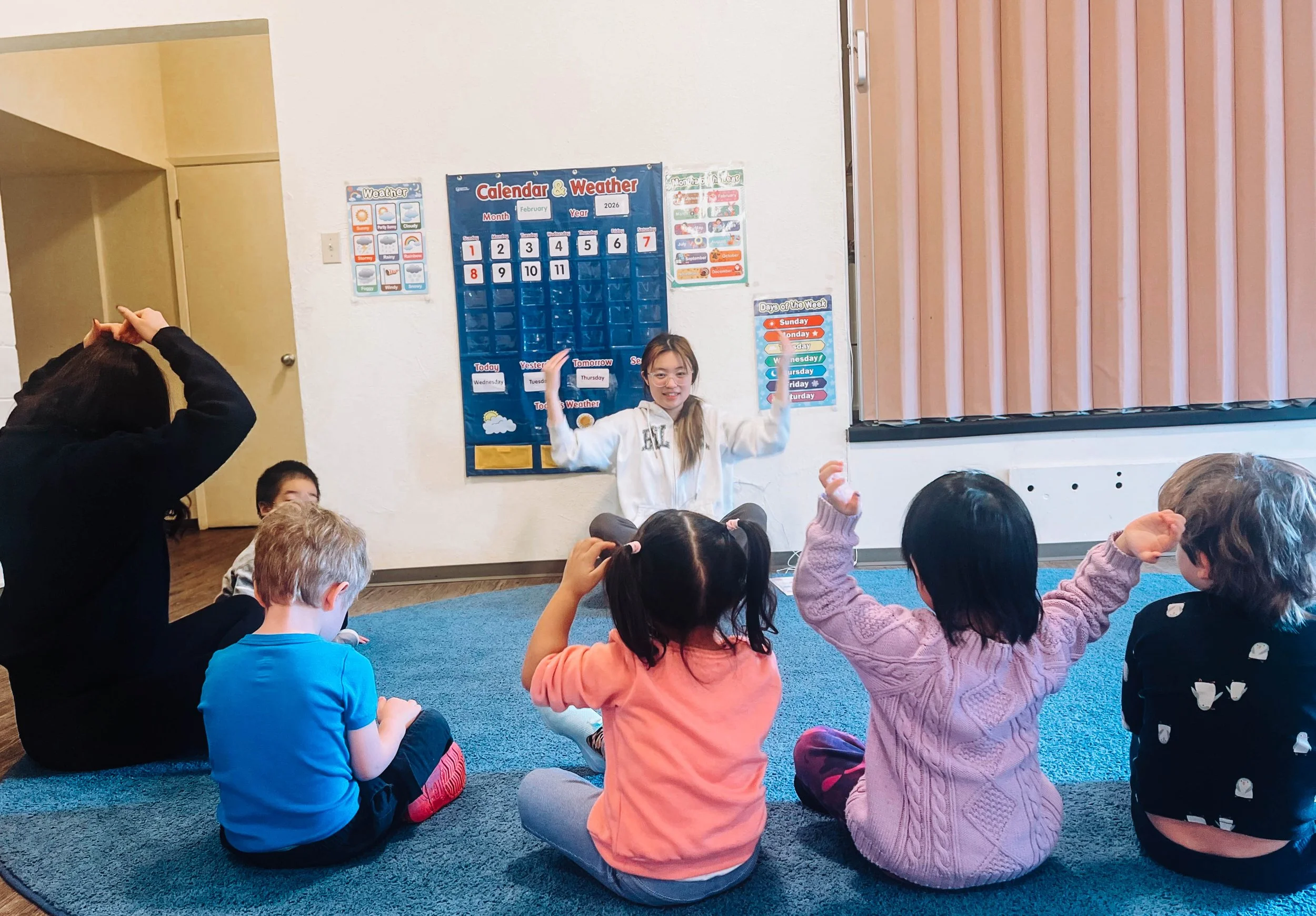 A group of young children and an adult sitting on a blue carpet in a classroom, with a girl standing in front of a calendar and weather chart, teaching or presenting to the others.