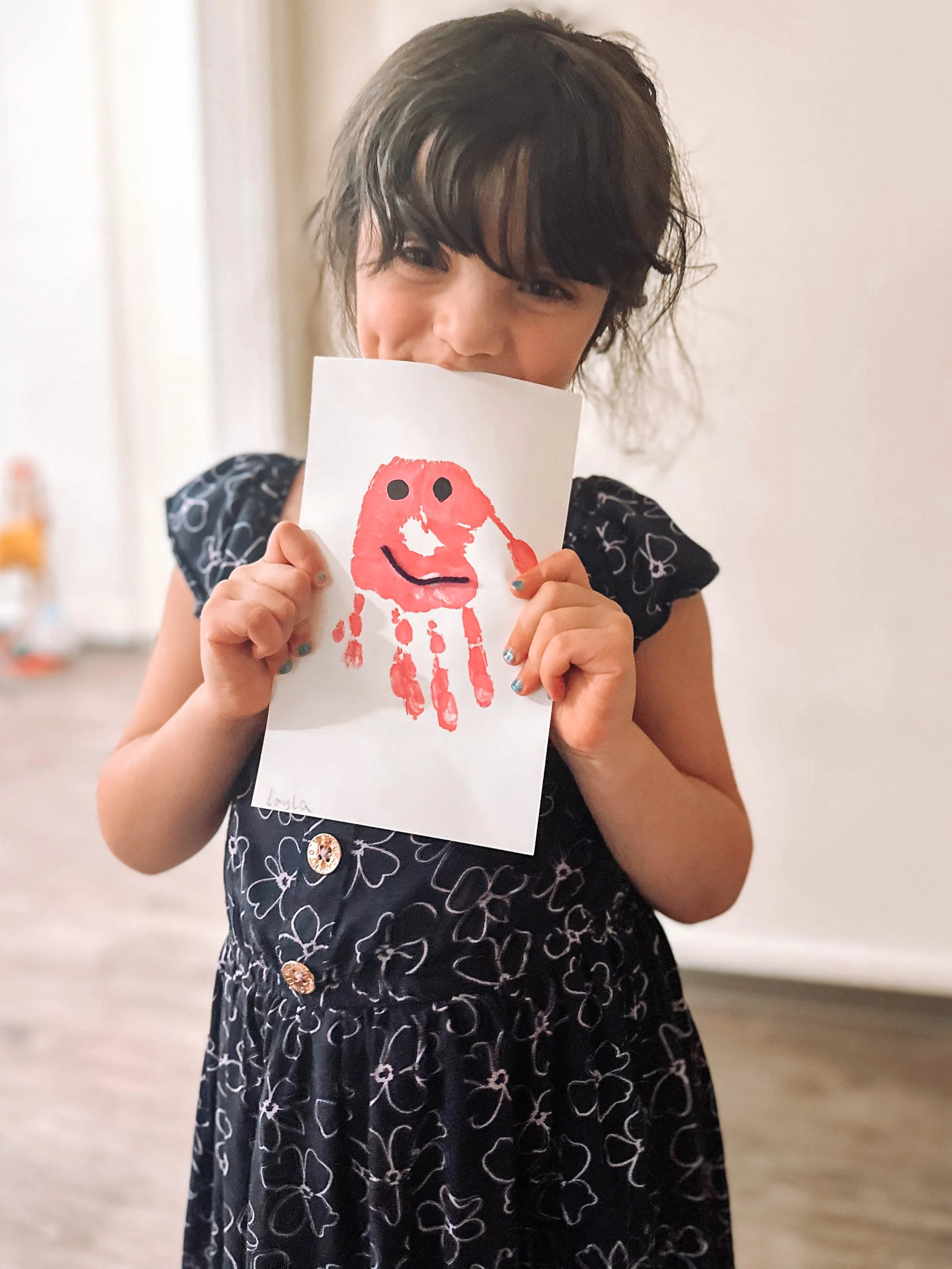 A young girl with dark hair, wearing a black dress with white floral patterns and buttons, is smiling while holding up a piece of paper with a red handprint and a smiling face painted on it.