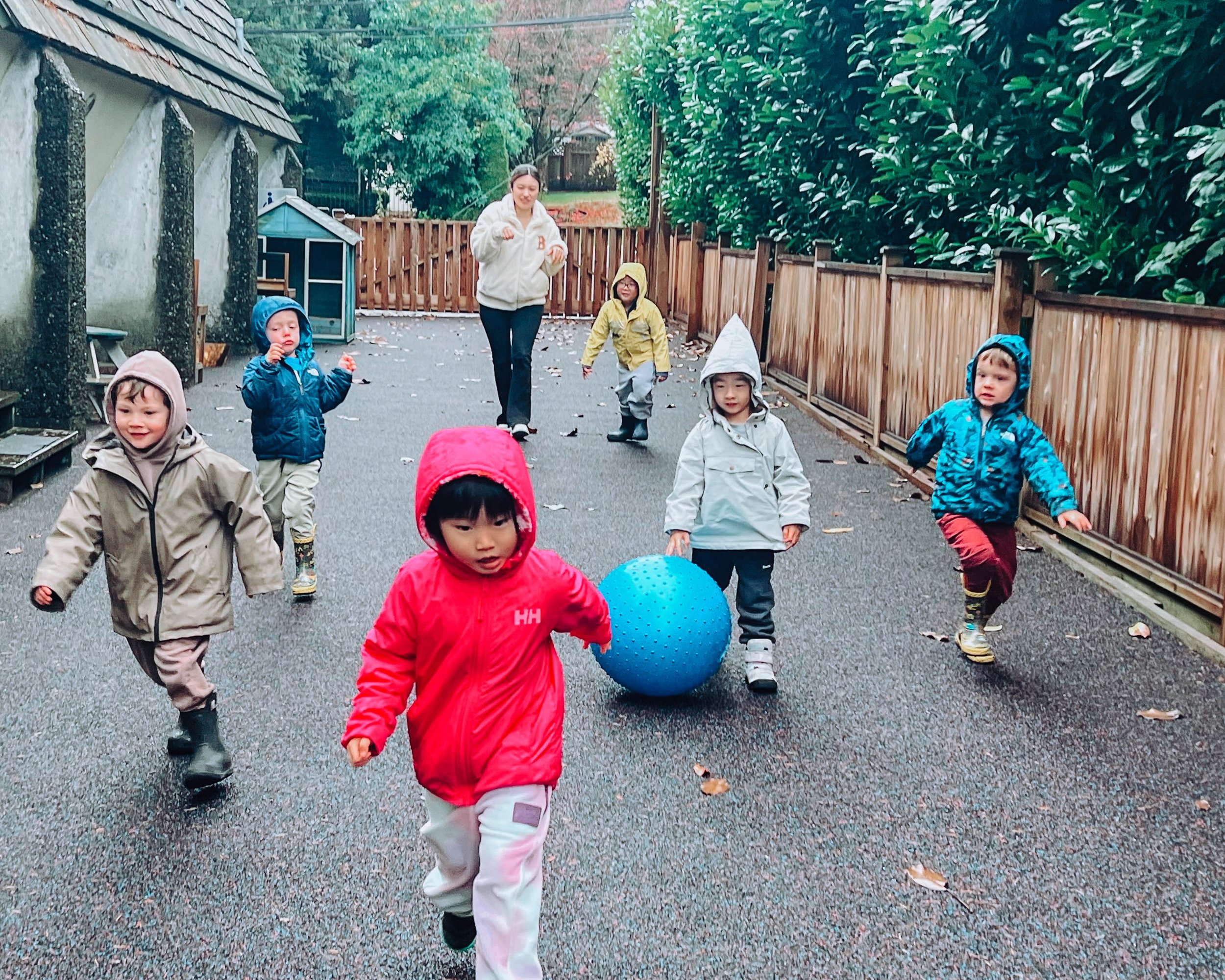 Children and a teacher playing outside in a wet, autumnal playground, with children wearing raincoats and boots, and a large blue ball on the ground.