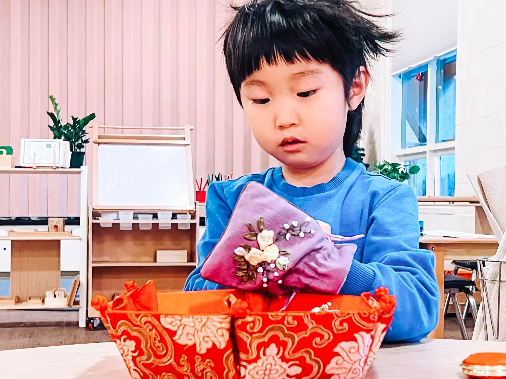 A young child with dark hair and wearing a blue shirt, looking at a decorated fabric item in a classroom or playroom with wooden shelves, plants, and large windows.
