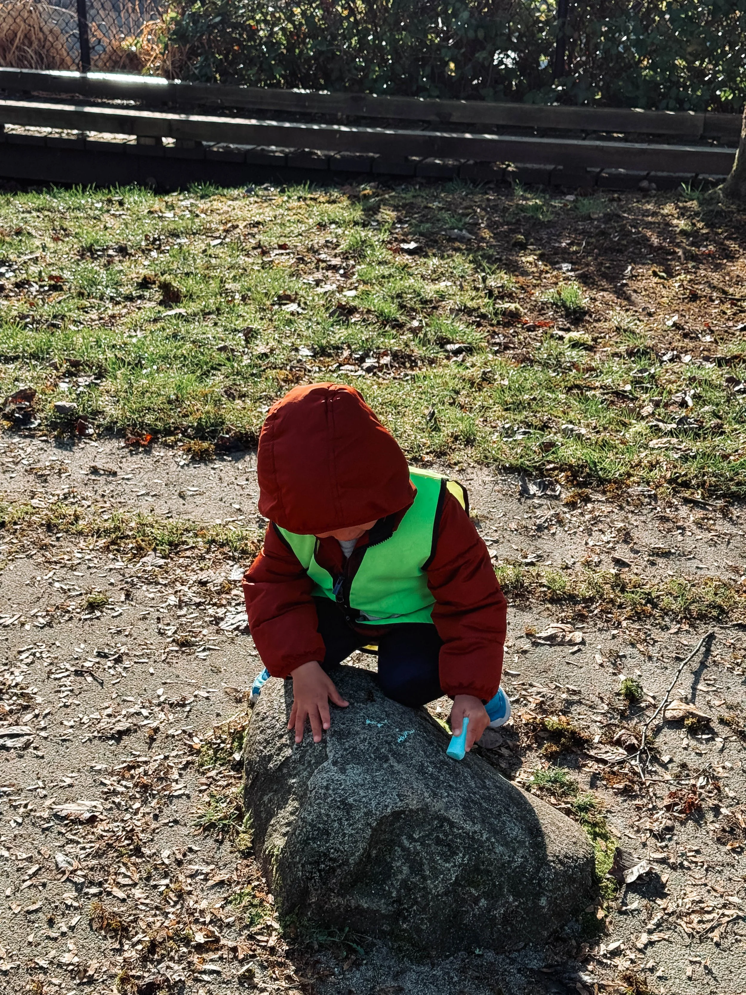 Young child wearing a red jacket and a neon green safety vest crouching on a rock outdoors, holding a chalk in his right hand, with grass and fallen leaves on the ground.