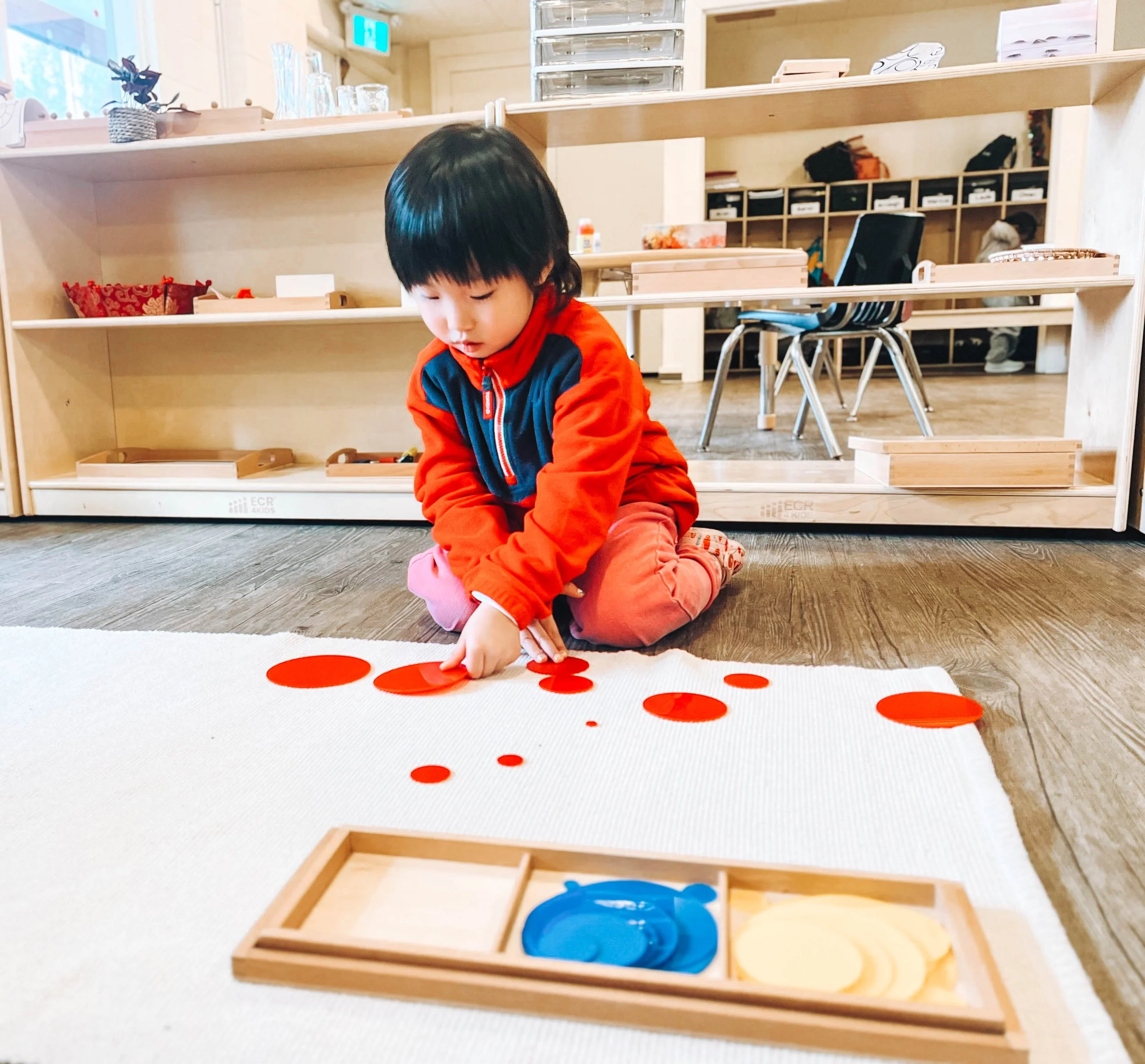 Young child in red and navy jacket playing with red circle stickers on a white rug in a classroom or playroom.