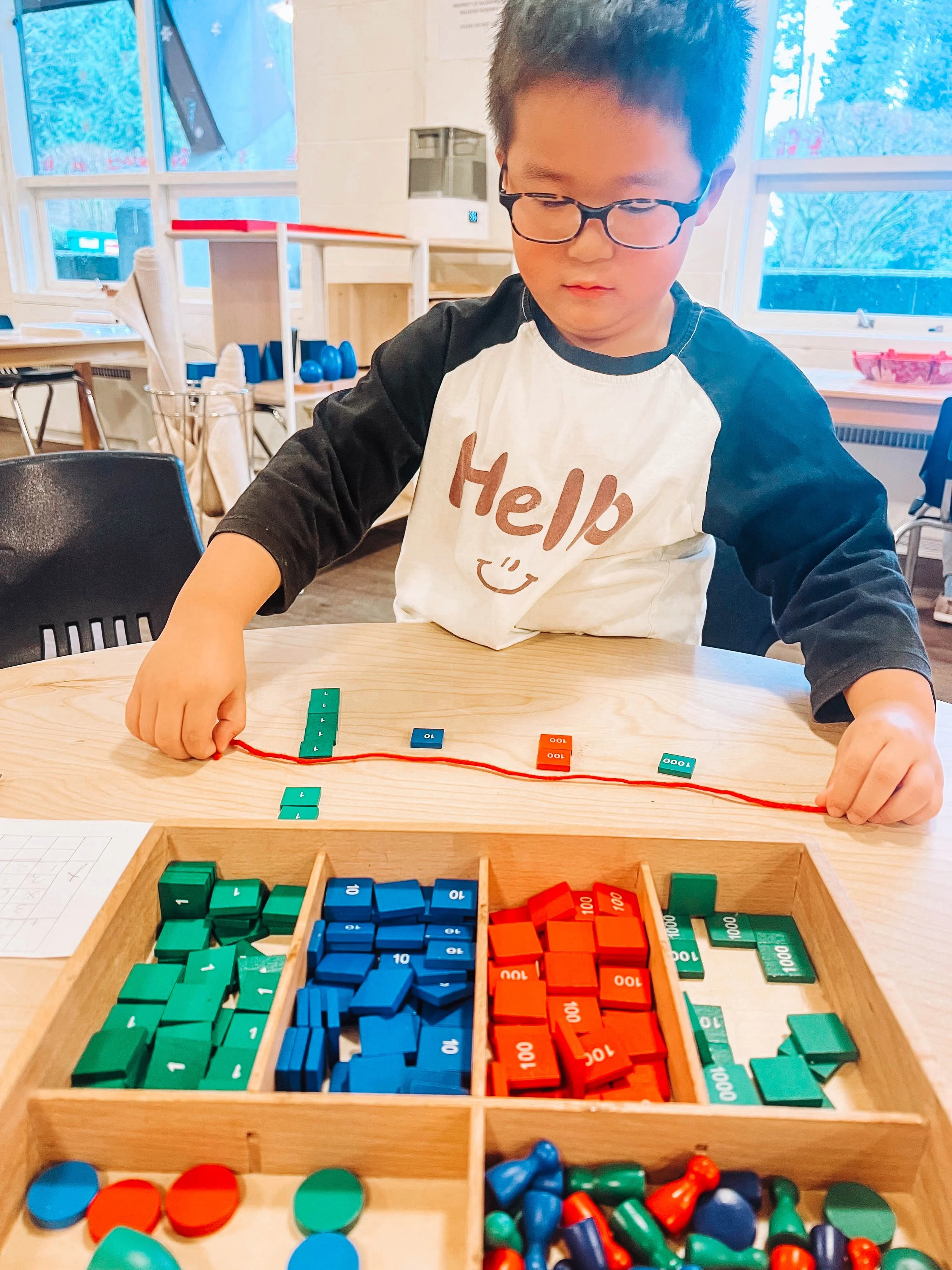 A woman with long hair taking a selfie in a tropical setting. She is wearing sunglasses and holding a phone, standing in front of pink flowers and lush greenery.
