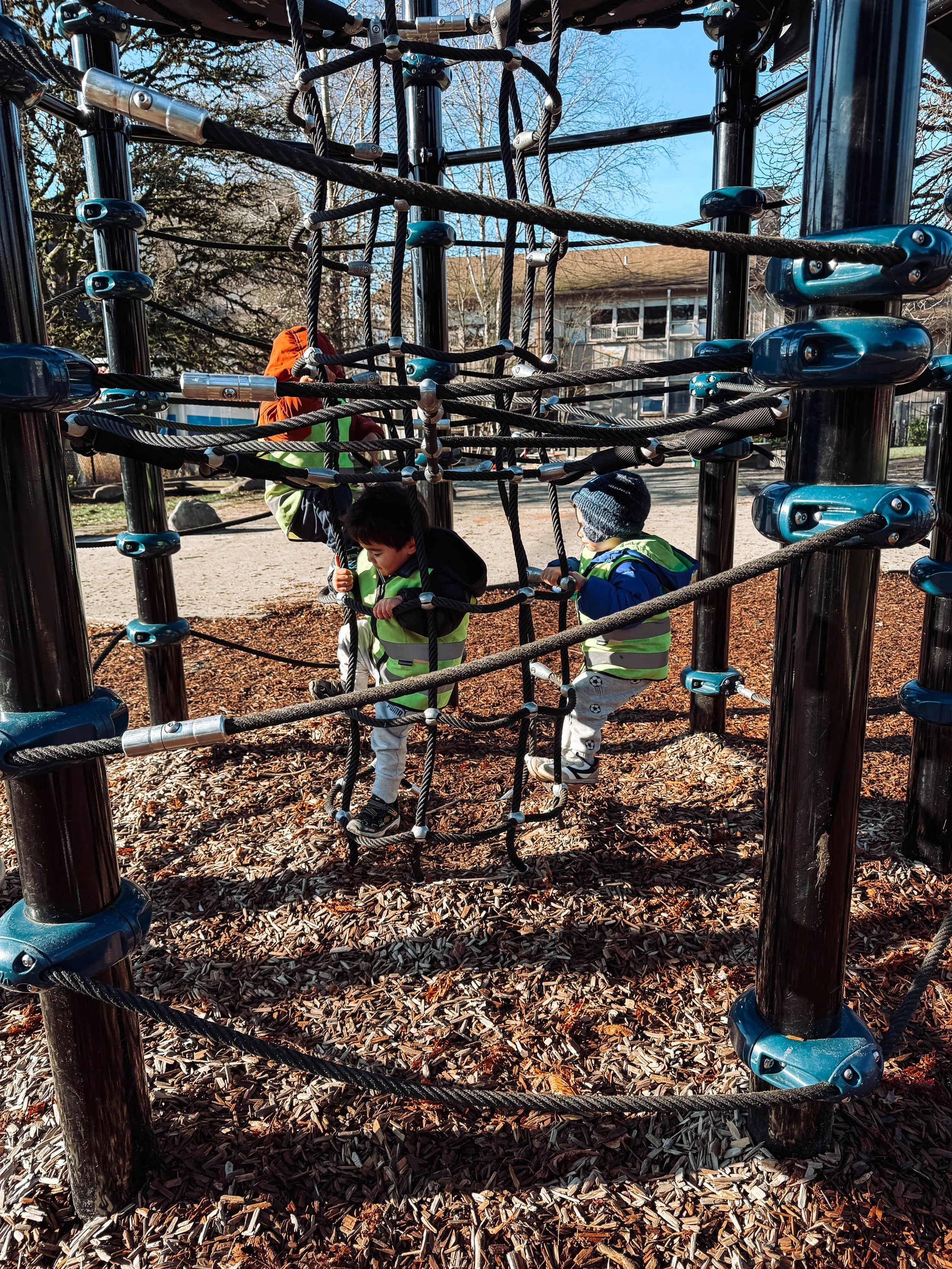 Three children playing on a jungle gym at a playground, with rope climbing elements, during a sunny day.