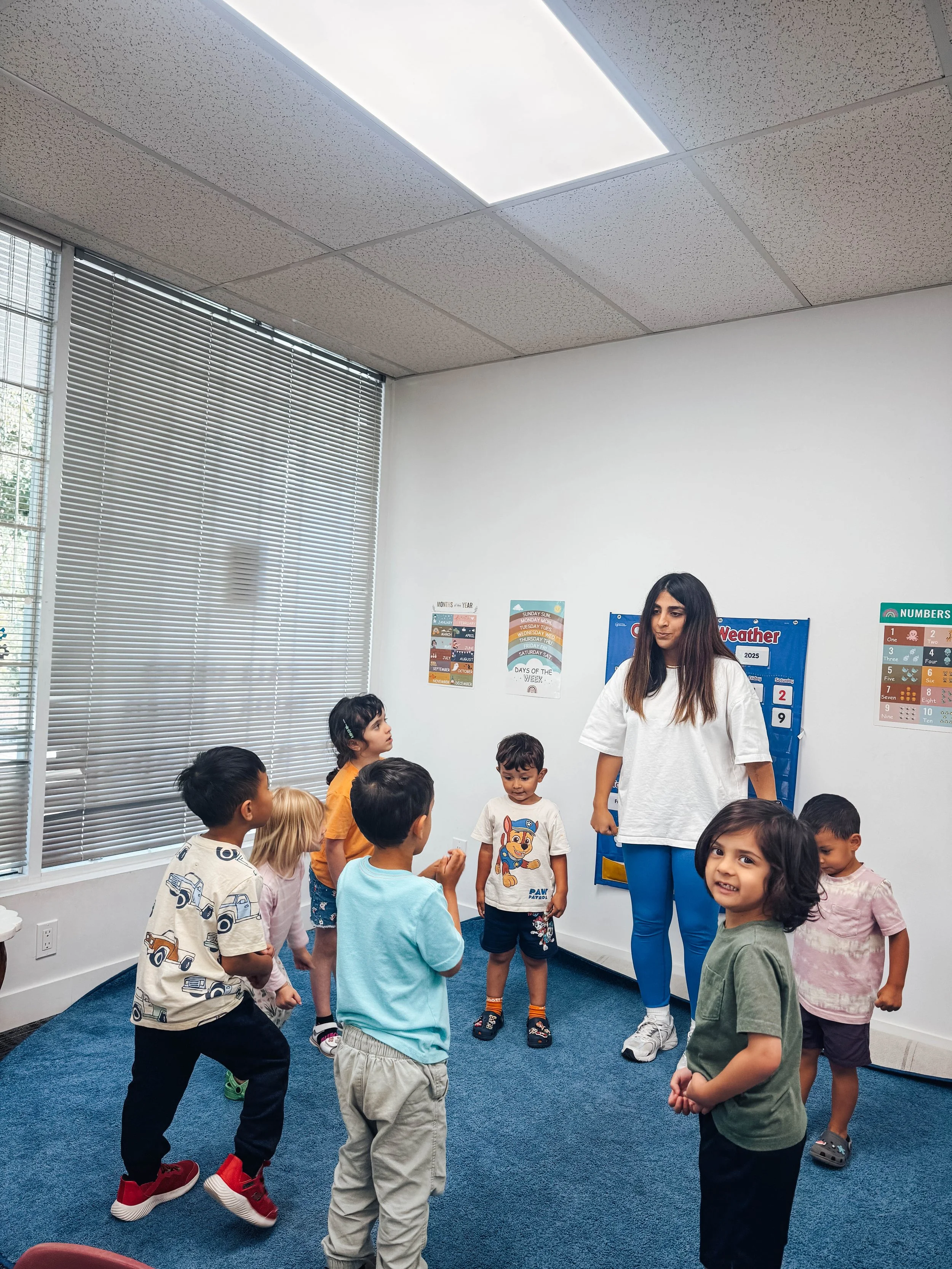A group of young children participating in a classroom activity led by a teacher, with educational posters on the white wall and window blinds in the background.