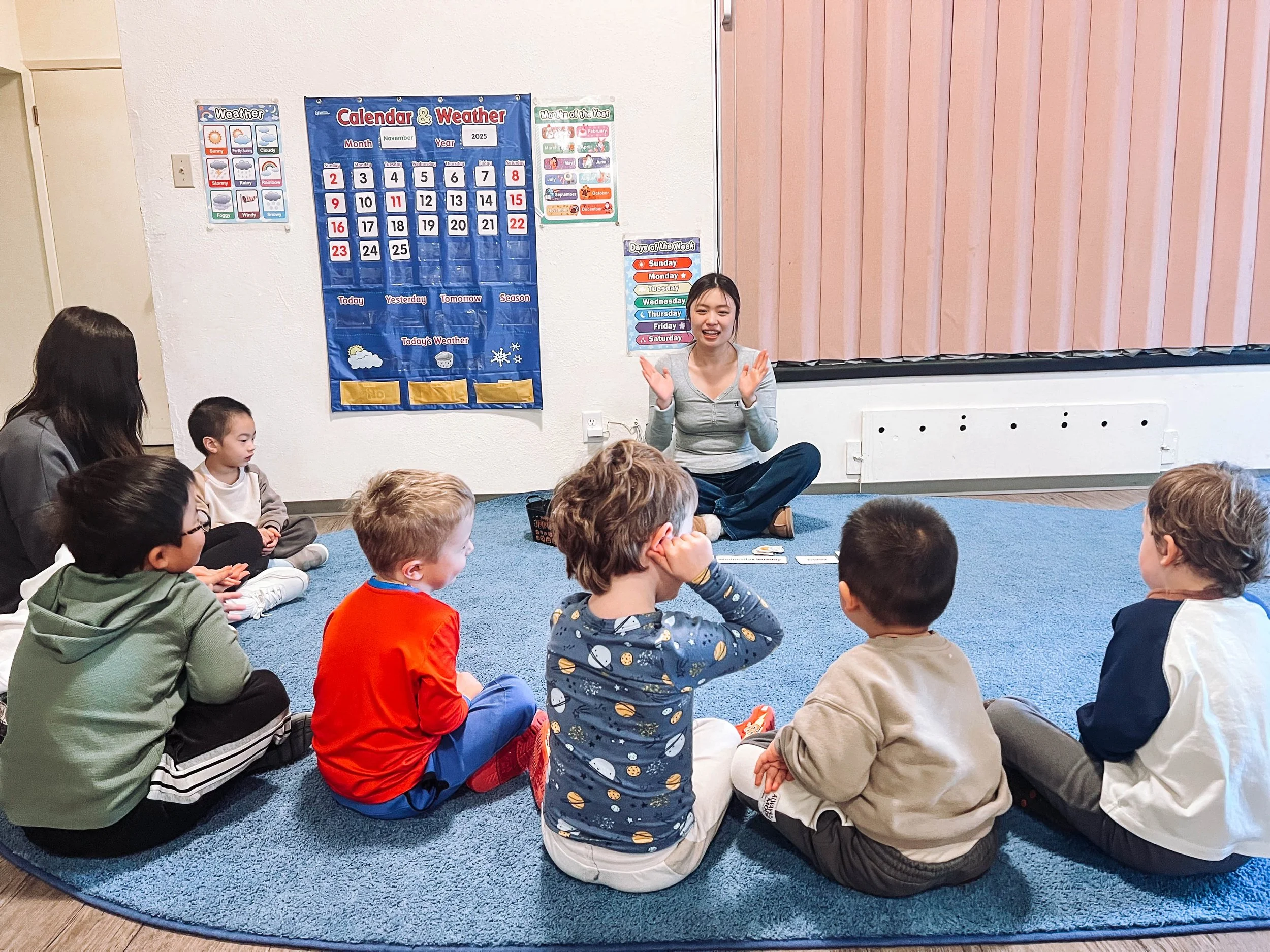 A teacher leading a classroom activity with young children sitting on a blue carpet in a circle, with educational posters on the wall.